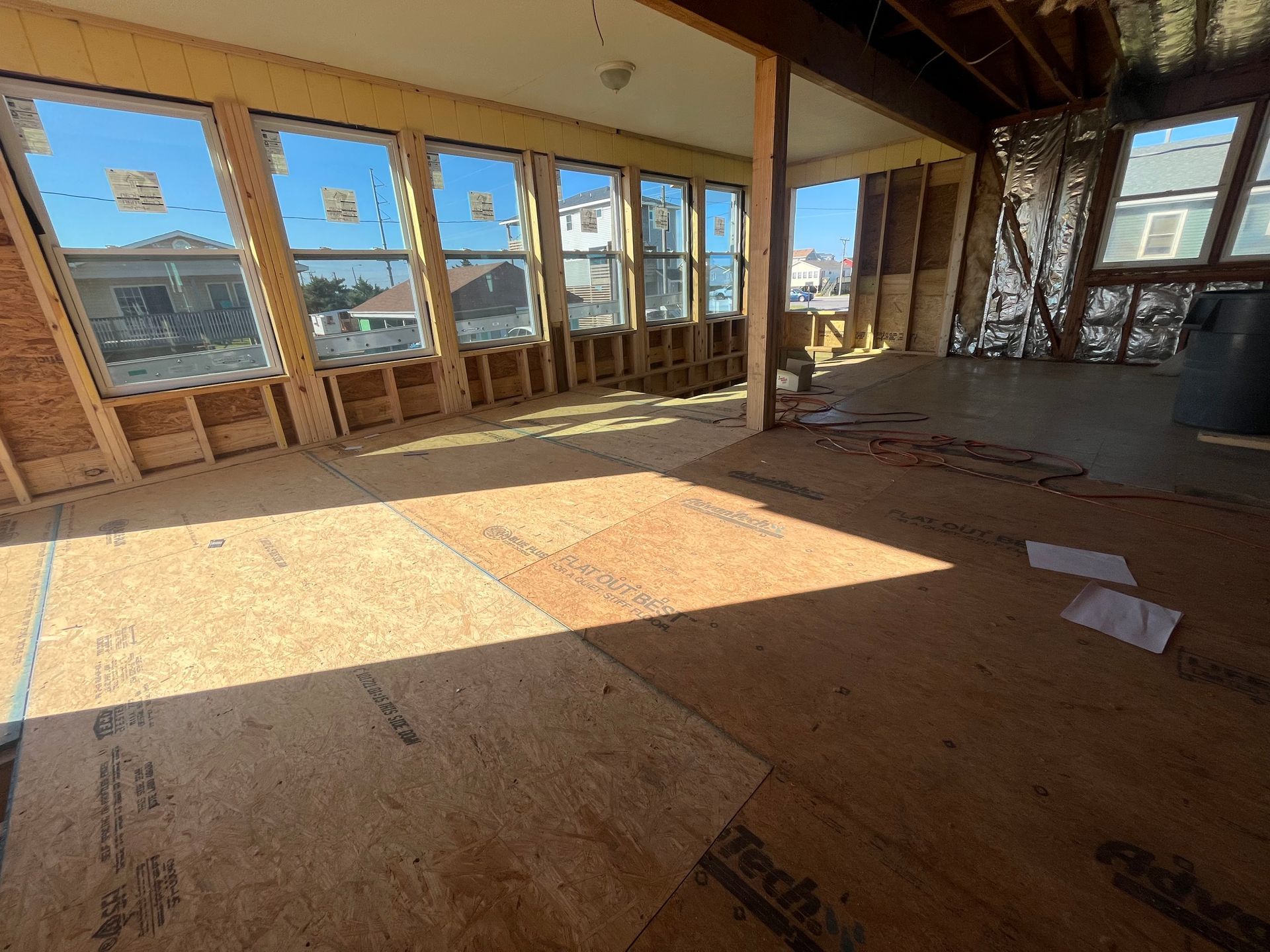 Interior view of a room under construction, with exposed wood framing, large windows, and unfinished floor.