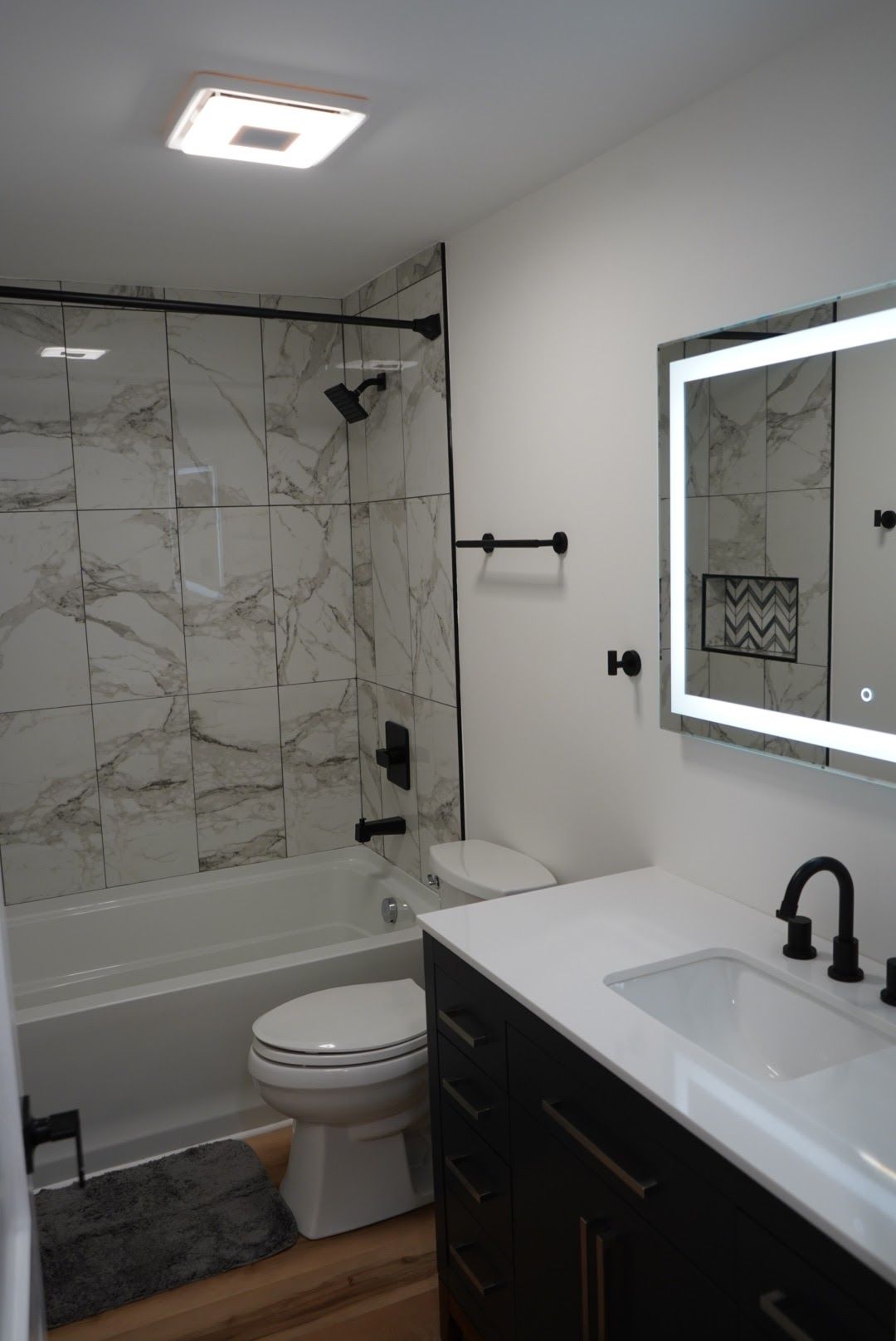 Modern bathroom with a white marble-tiled shower, black fixtures, a dark vanity, and a lighted mirror.