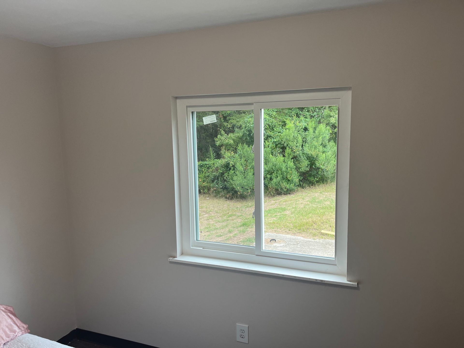 A room with a white-framed window overlooking a green outdoor area. Beige walls and a light-colored window sill.
