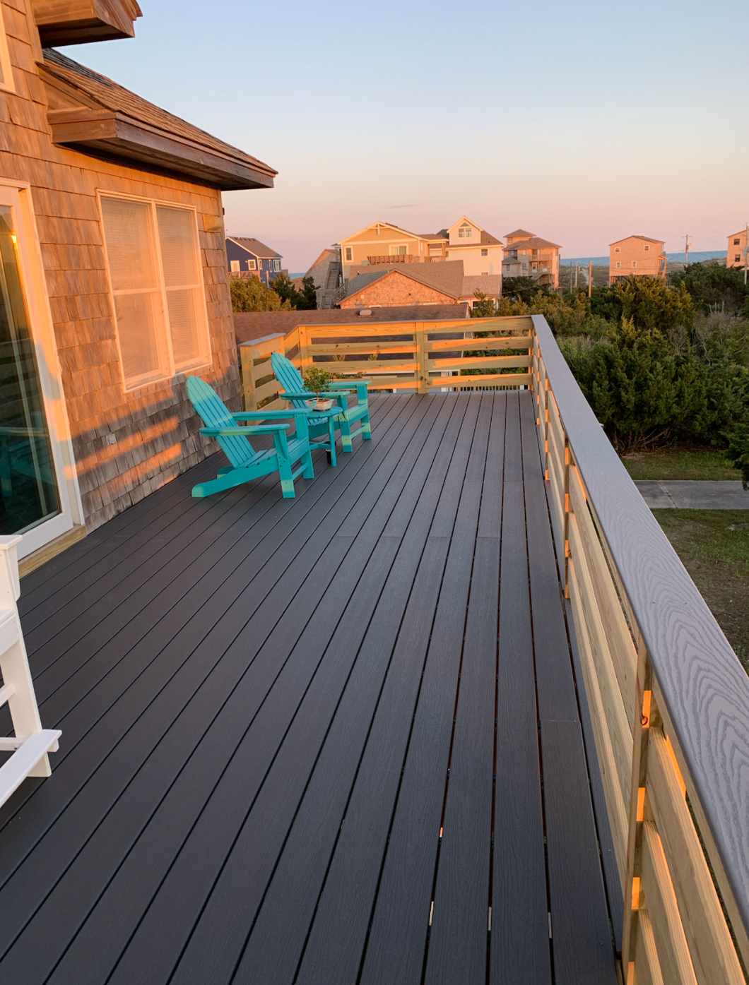 Wooden deck with blue Adirondack chairs at sunset, overlooking houses and greenery.