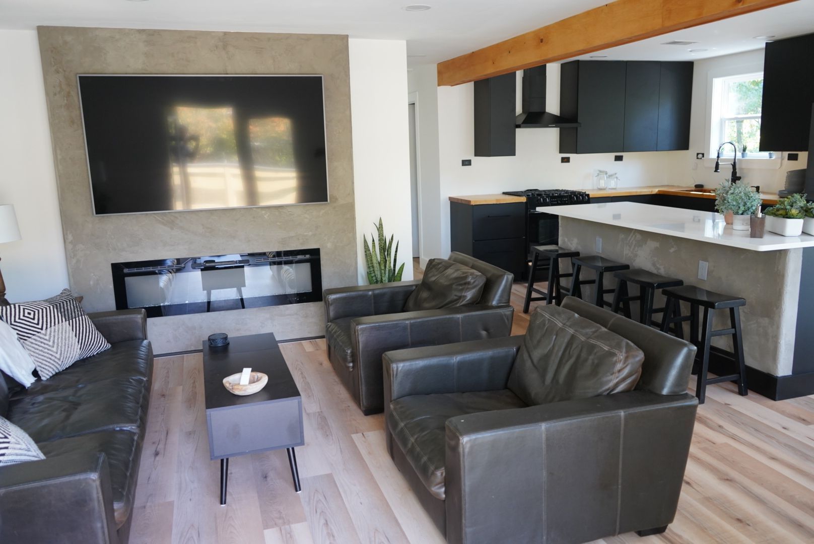 A modern living room with a black leather sofa and armchairs, a fireplace, and an open kitchen with black cabinets.