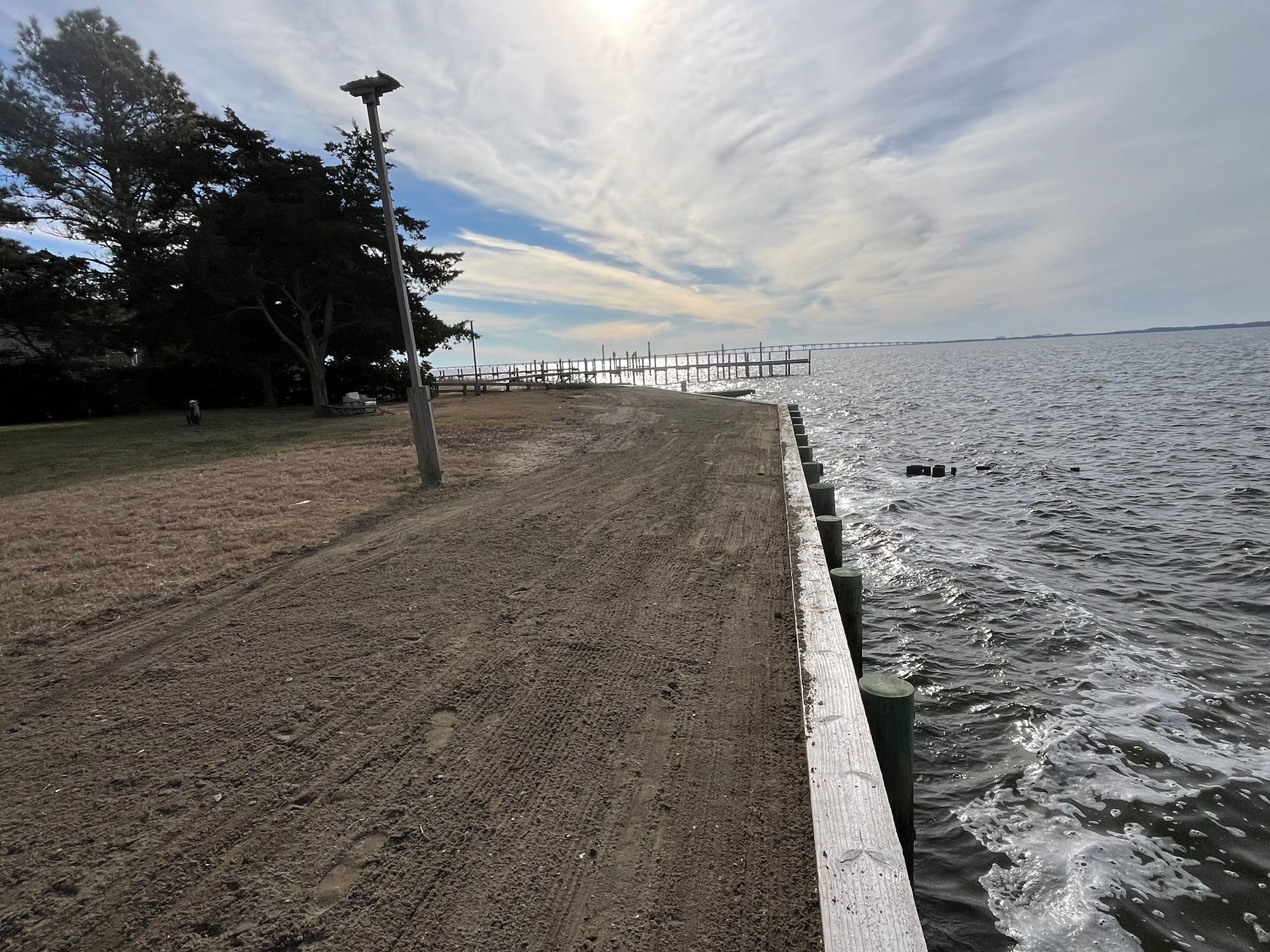 A dirt path alongside a body of water on a sunny day, with a wooden border and light post.
