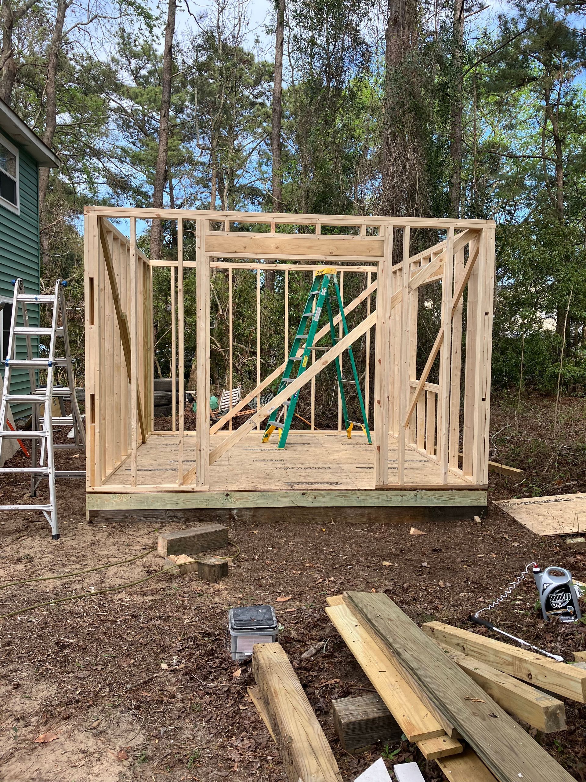 Wooden frame of a shed under construction outdoors, surrounded by trees and tools.