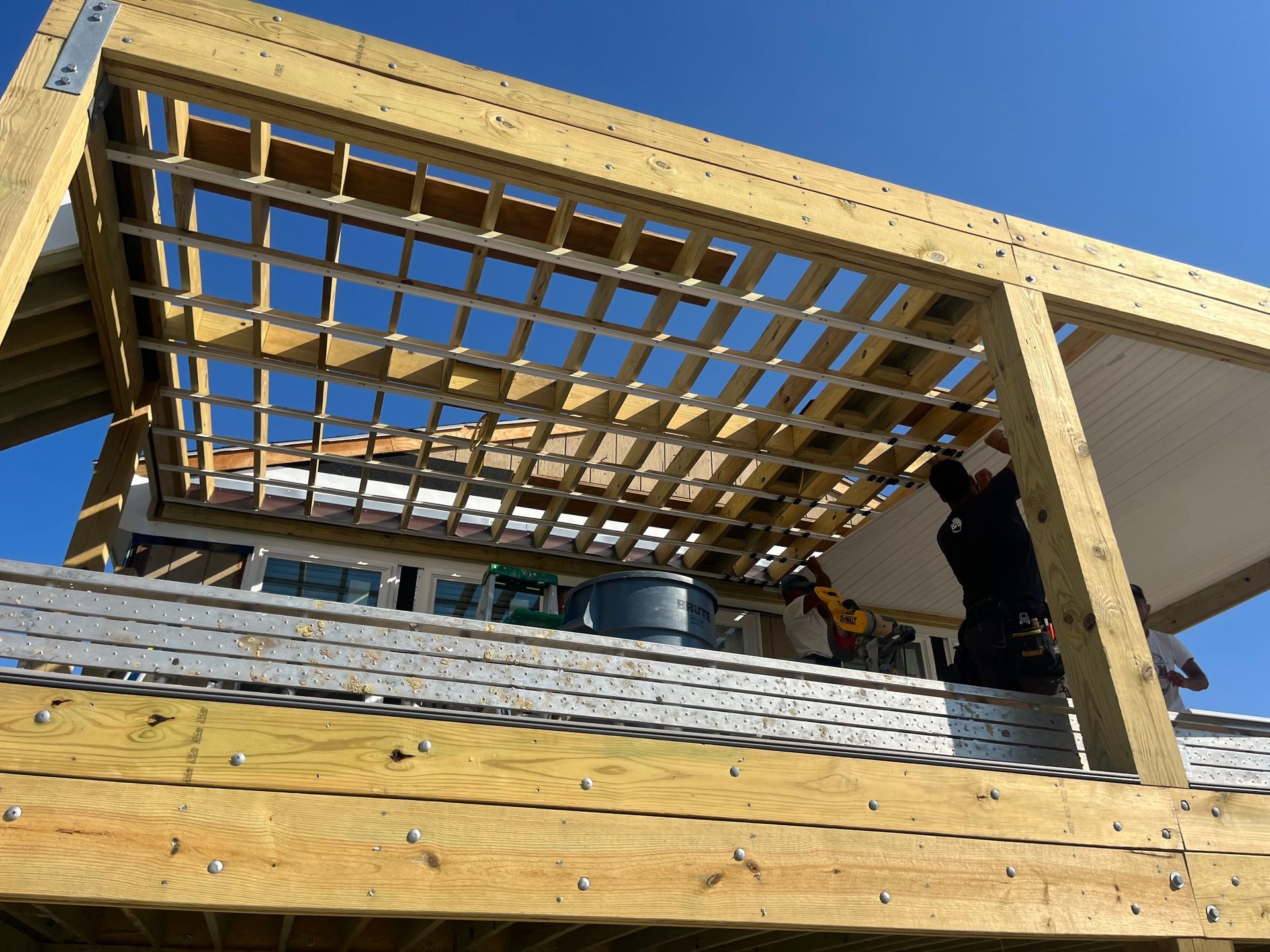 Construction of a wooden pergola over a roof deck with a worker installing the structure. Bright blue sky.