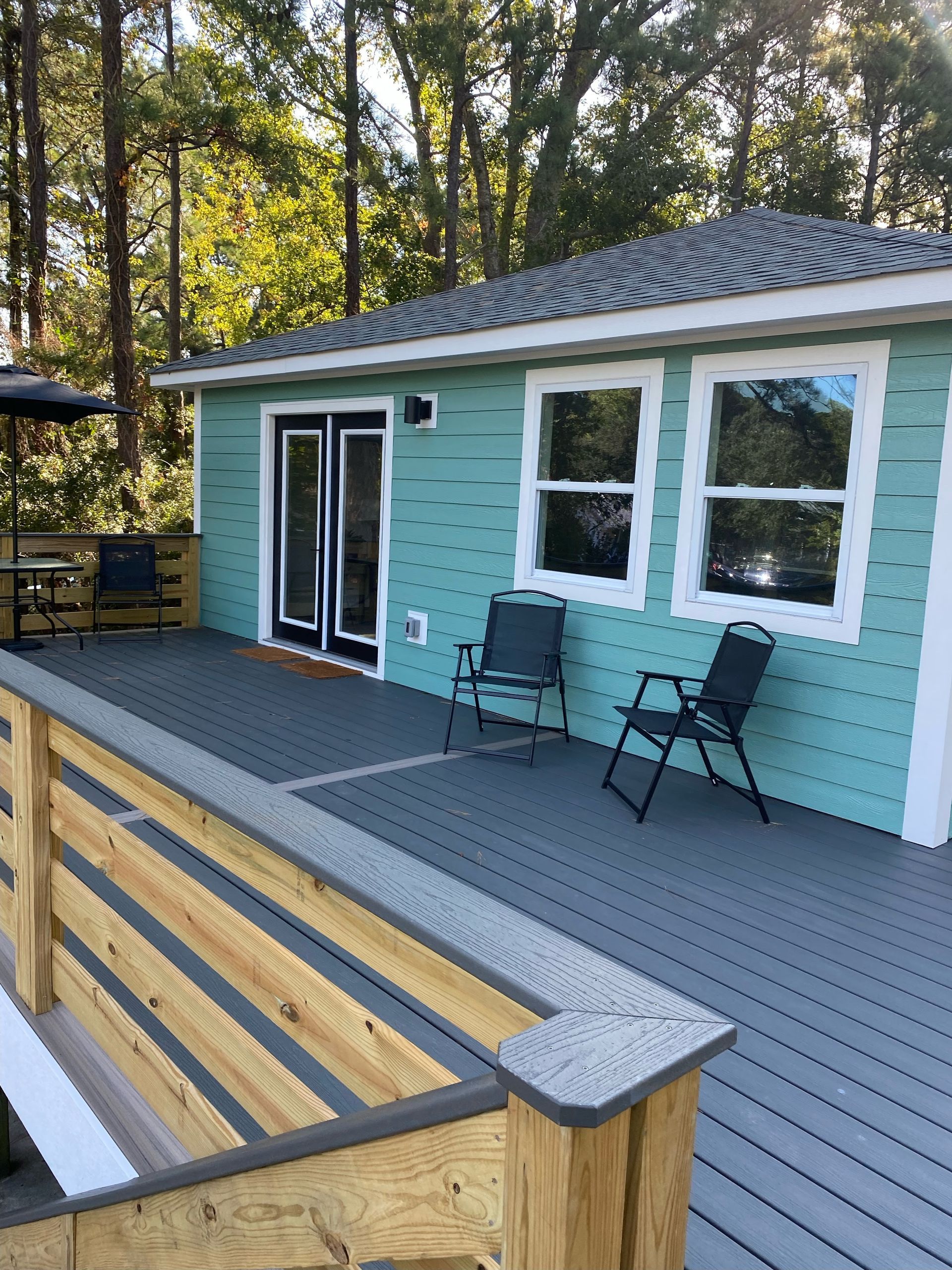 A small teal house with a gray deck and two black chairs. French doors and windows line the front, set in a wooded area.