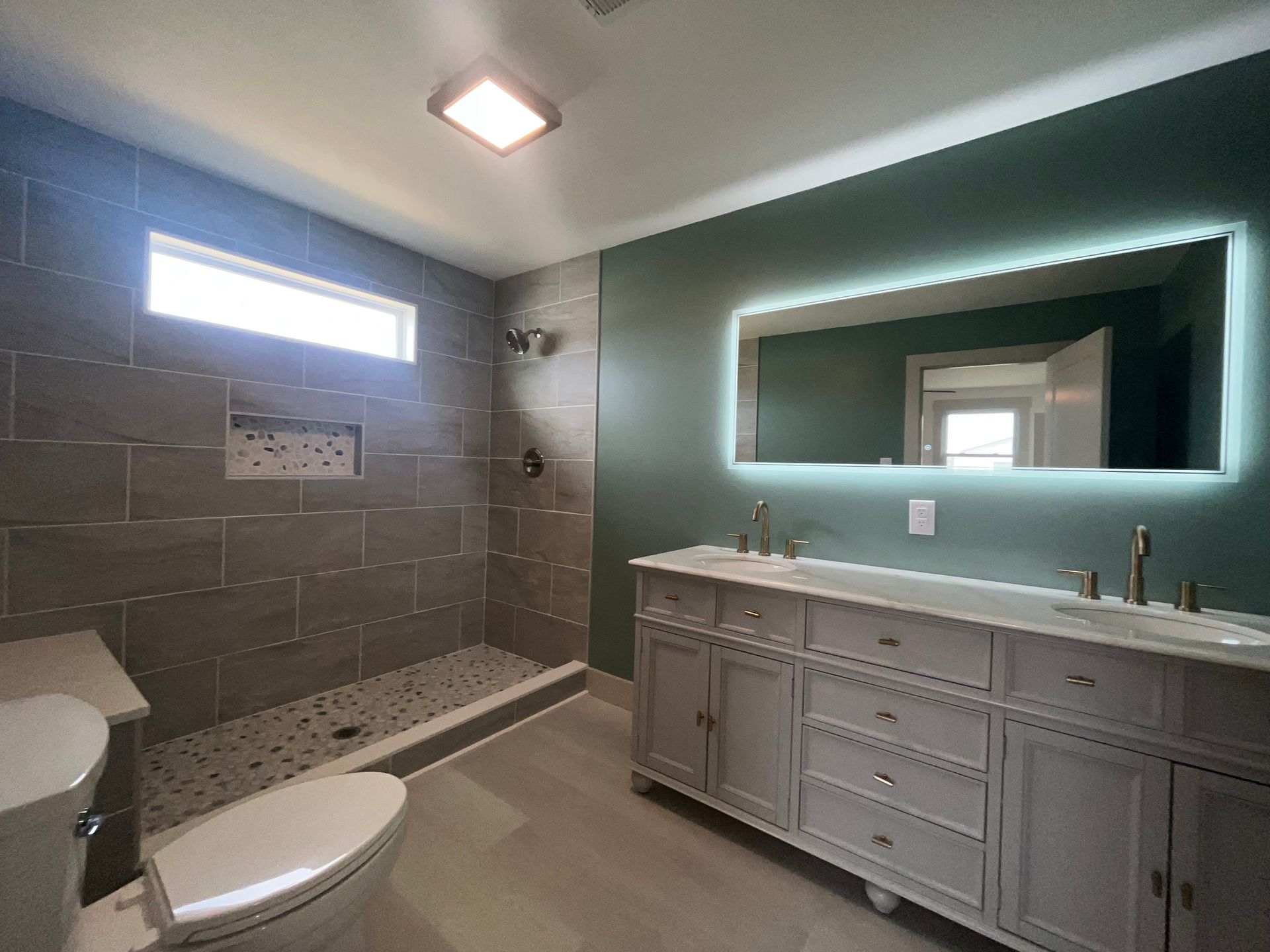 Modern bathroom with gray tiled shower, toilet, vanity, and green wall with illuminated mirror.