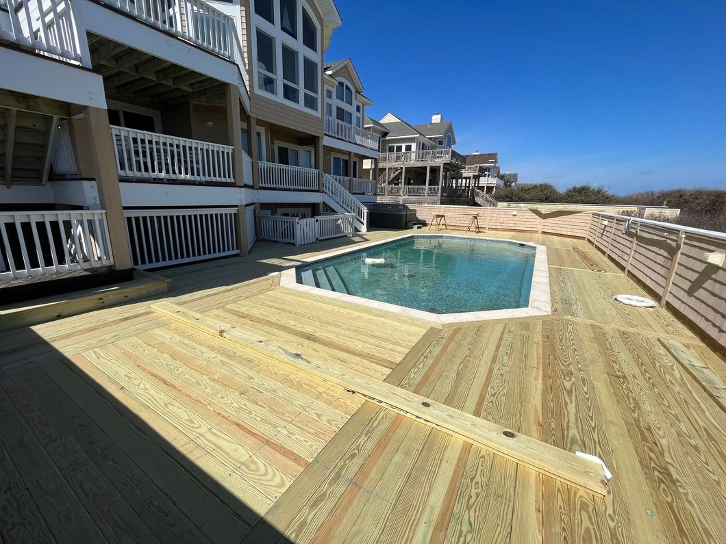 Wooden deck with a pool, overlooking the ocean. Large beach house in the background.