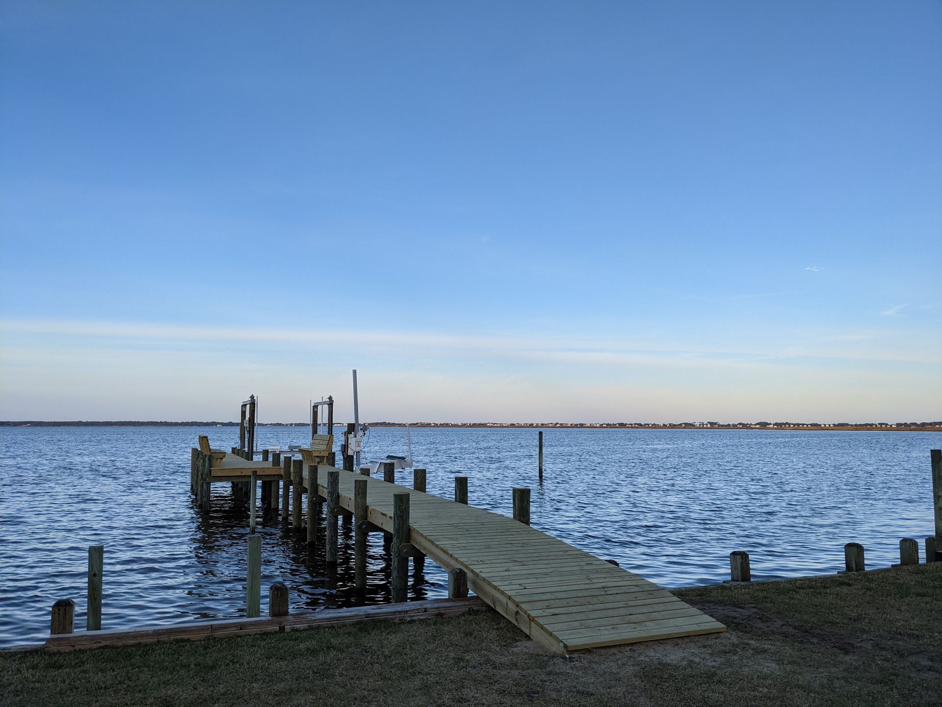 Dock extending into a body of water under a blue sky. The dock has a ramp and is surrounded by water and a grassy bank.