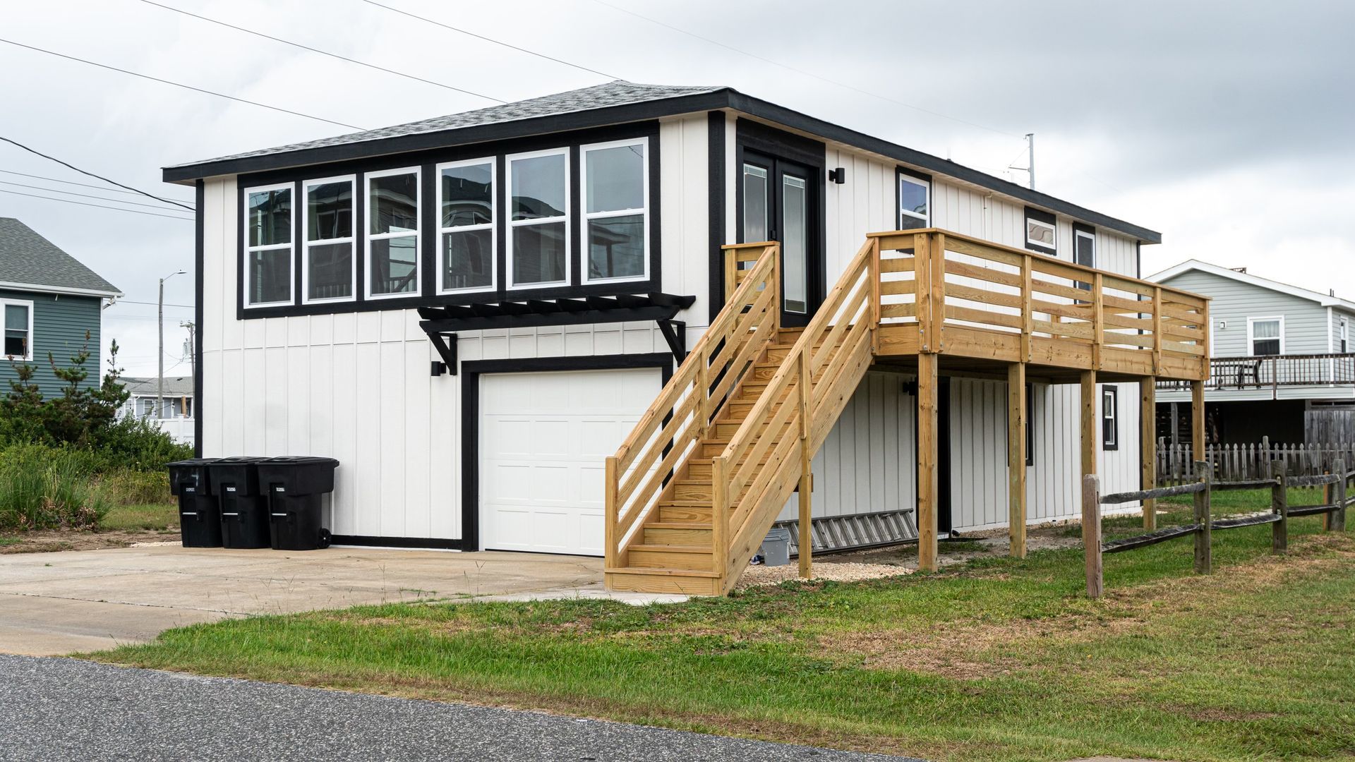 Two-story white house with black trim and a wooden deck and stairs. Three black trash bins sit on the concrete driveway.