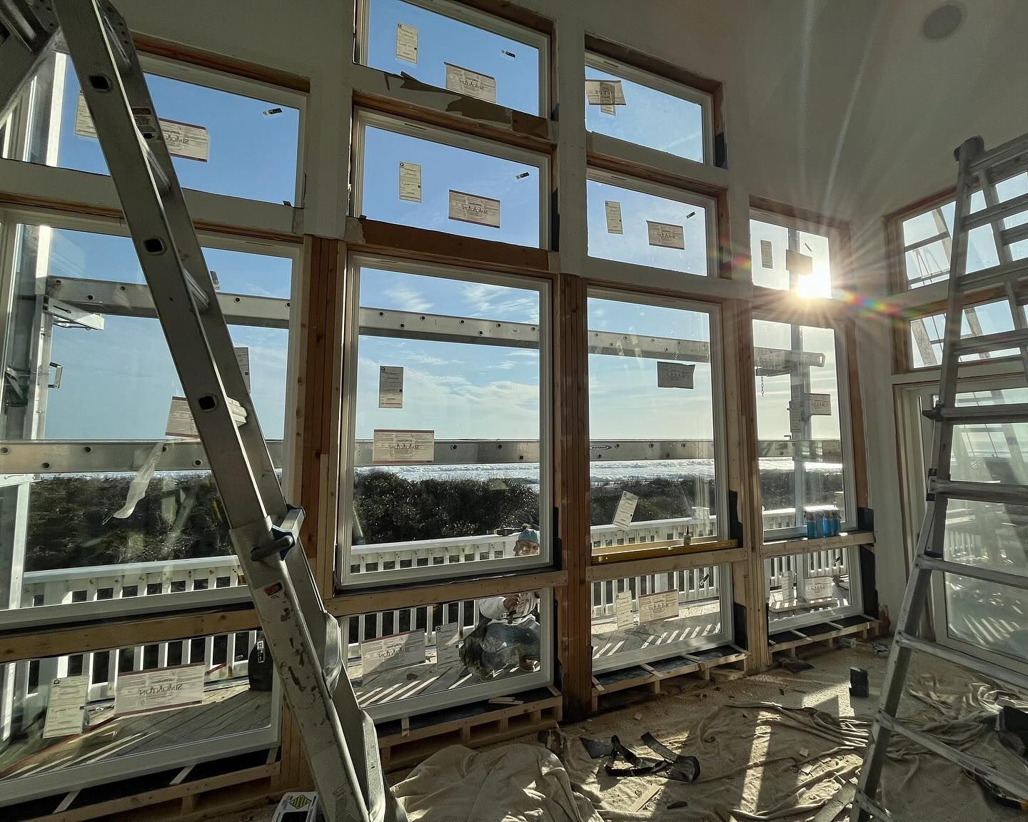 Interior view of a room with large windows during construction, sunlight streams in. A ladder leans against the window frame.