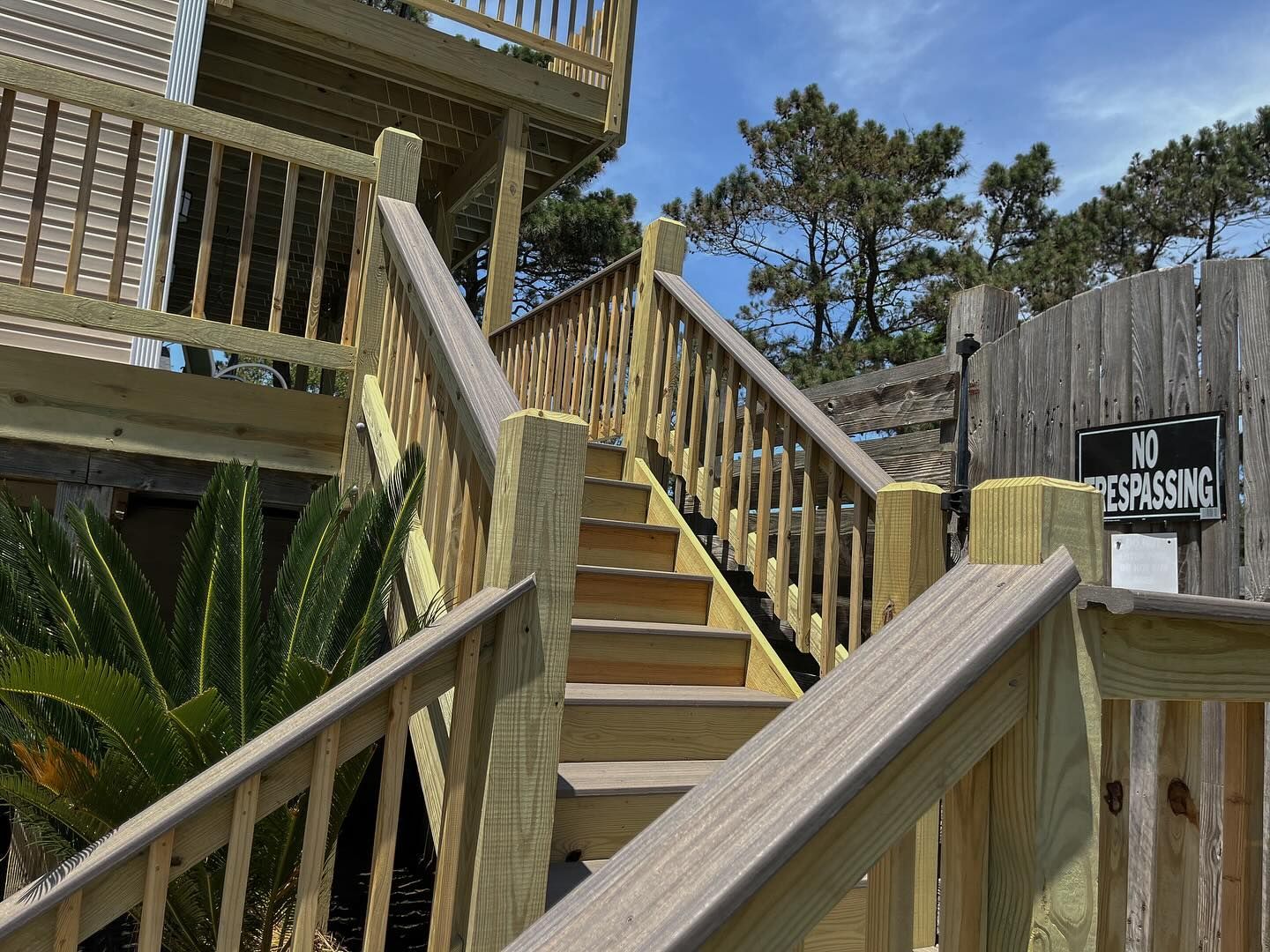 Wooden outdoor staircase leading up to a deck, with railings and steps. Bright sunlight and blue sky in the background.