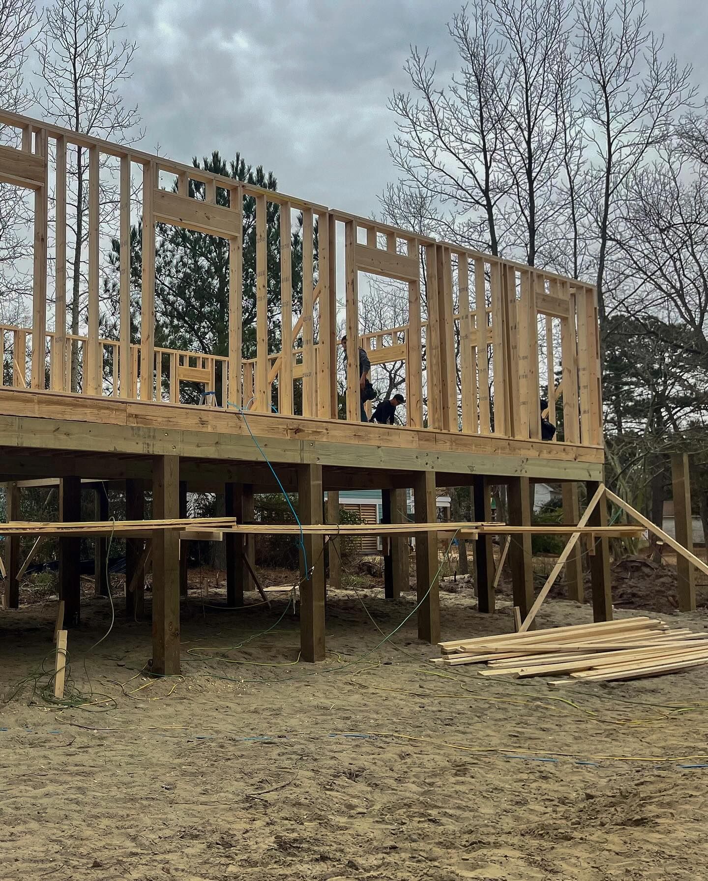 Wooden frame of a house under construction on elevated supports. Cloudy sky, bare trees, and dirt ground.