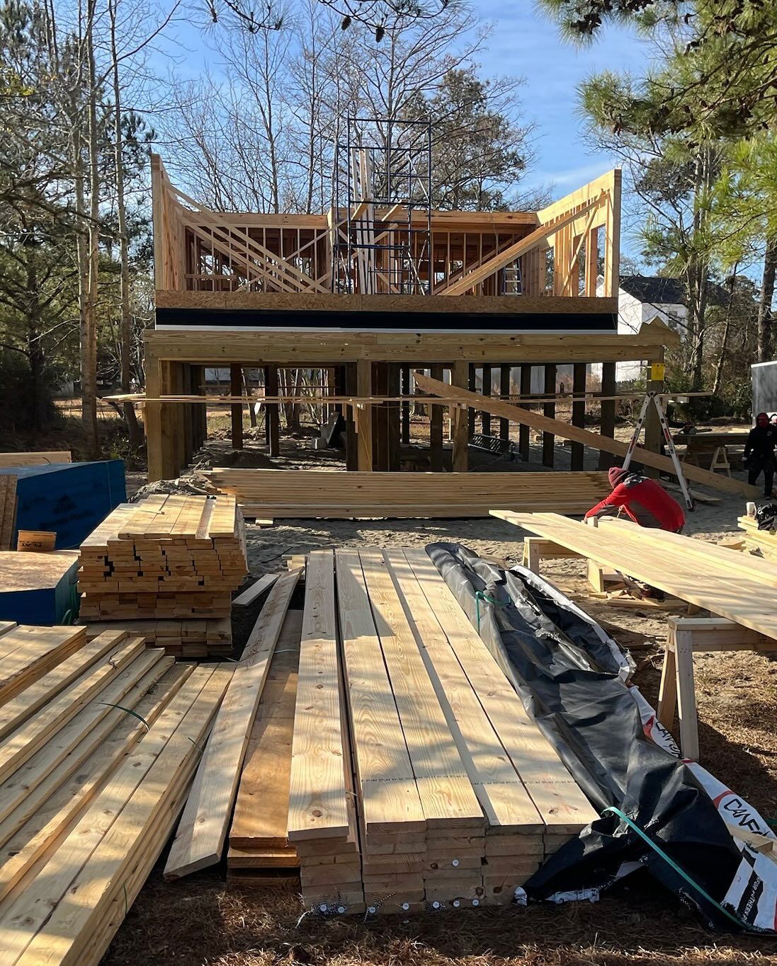 Construction of a two-story wood frame building on stilts; lumber piles and workers in the foreground.