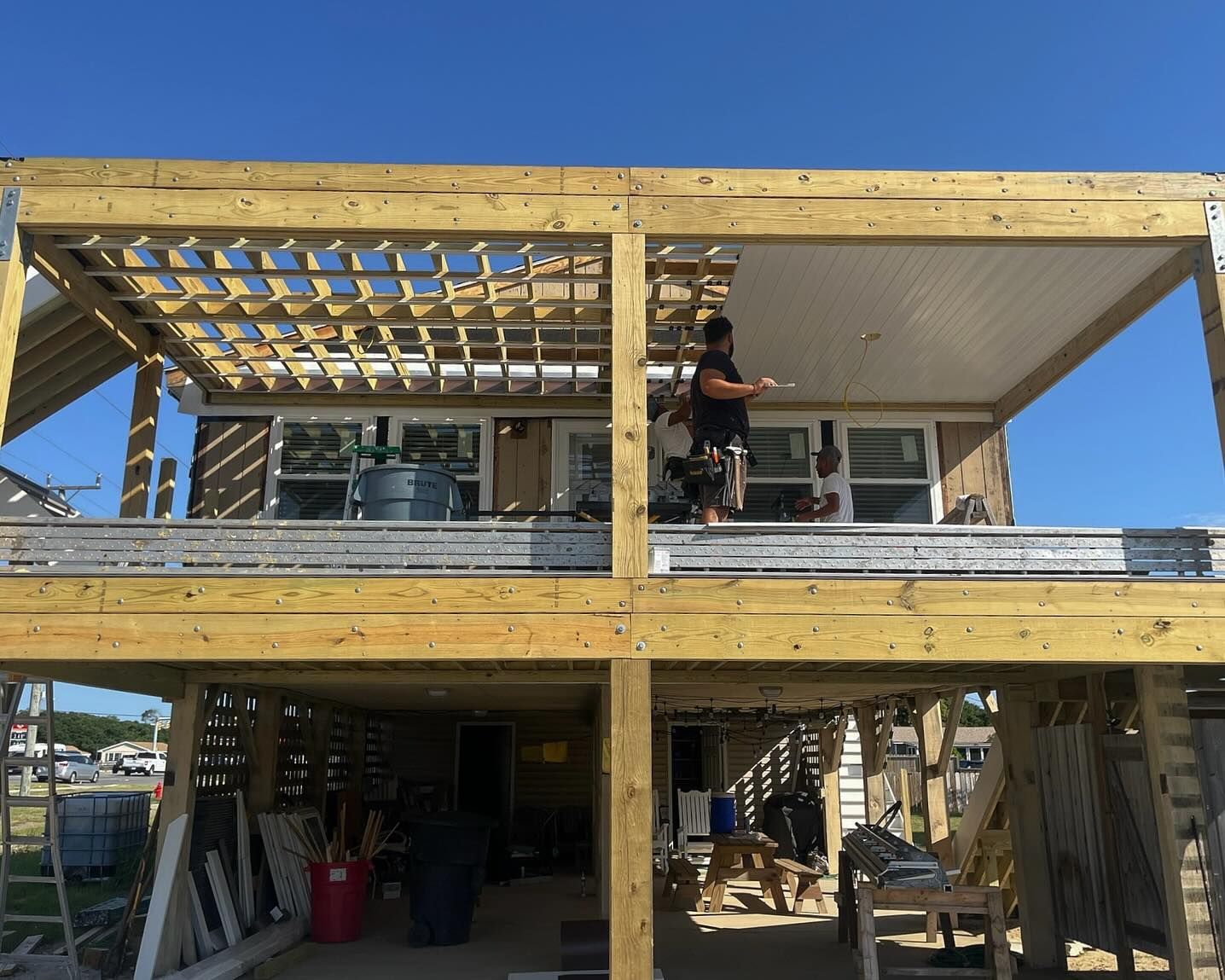 A person paints the ceiling of a wooden deck with a latticed section, overlooking a lower deck and beach setting.