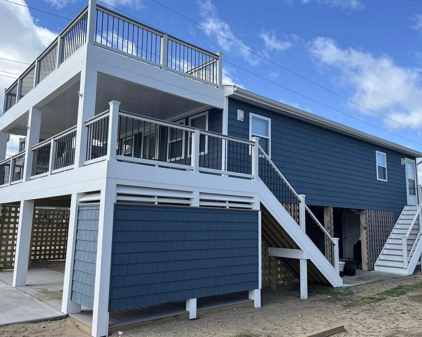 Two-story blue house with white railings and stairs.  Upper balcony overlooks a sandy area under a partly cloudy sky.