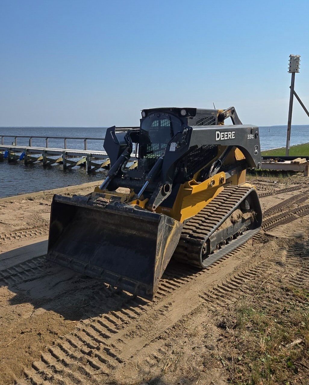 A yellow and black skid steer with a bucket on a sandy beach near a dock on a sunny day.