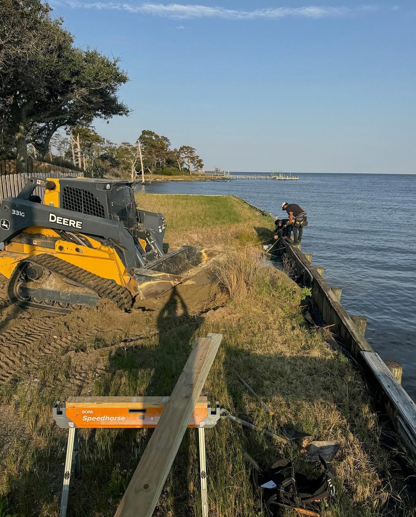 Construction site on a waterfront: A person works on a wooden barrier while a small bulldozer sits nearby.