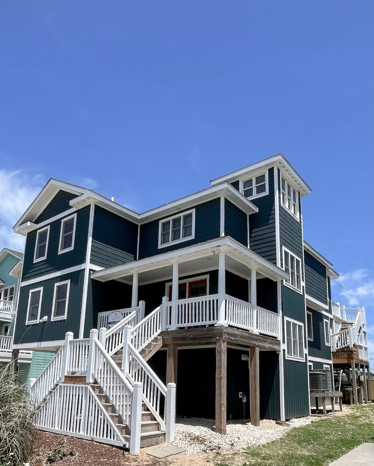 Dark teal beach house with white trim and porch, under a bright blue sky. Staircase leads to the porch.