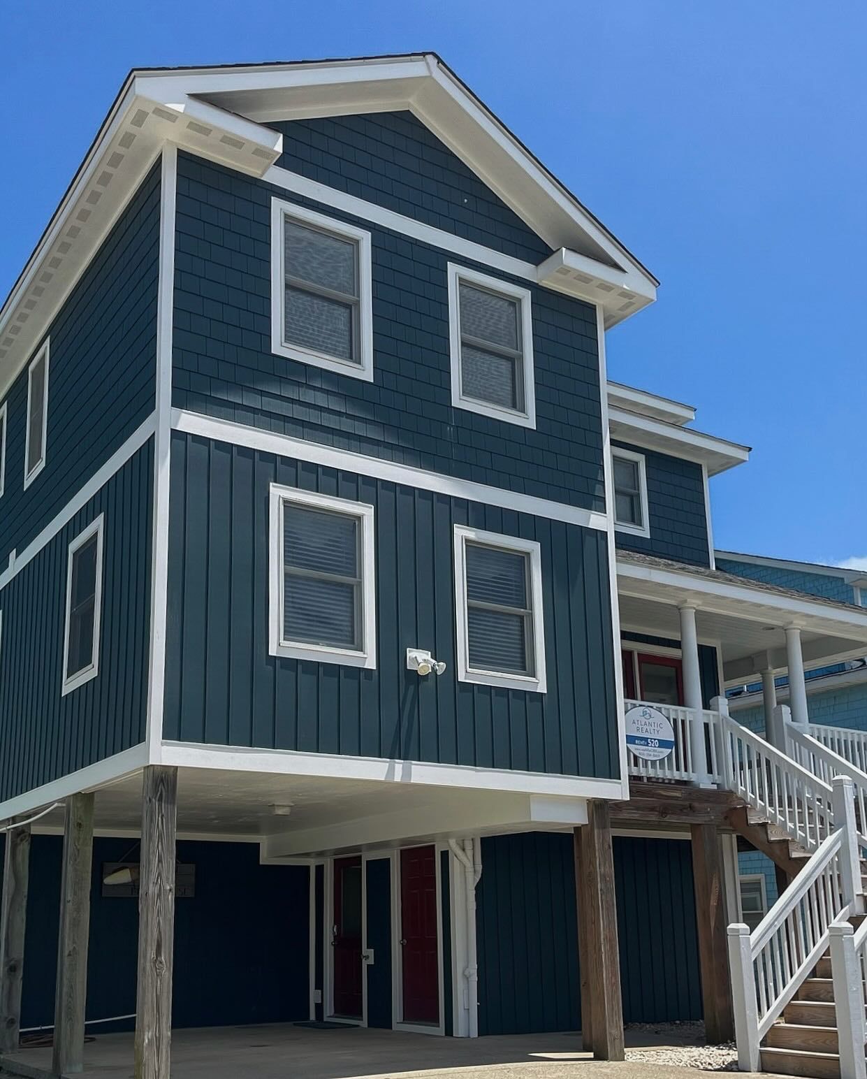 Two-story blue house on stilts with white trim and windows against a blue sky. Stairs lead up to a porch on the right.