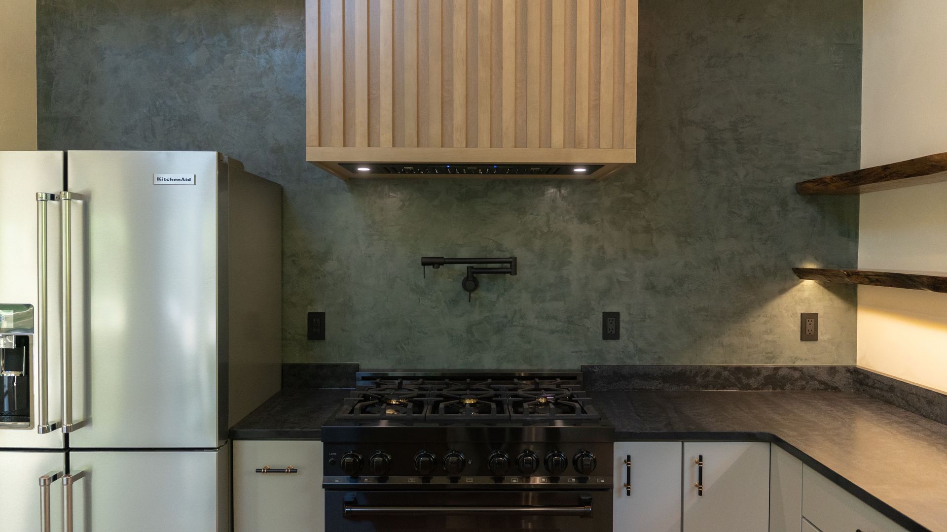 Kitchen with stainless steel refrigerator, black range, green textured backsplash, and wooden range hood.