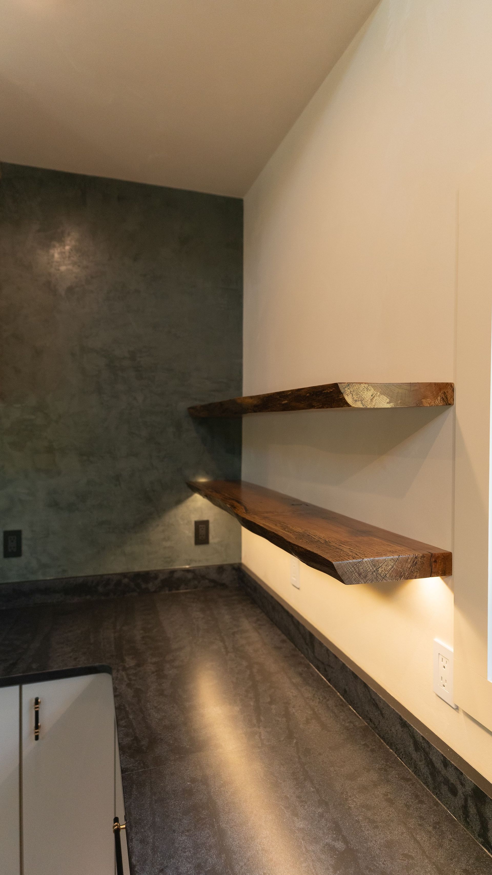 Two rustic wooden shelves on a cream wall, above a gray countertop with cabinet below. Dark green textured wall behind.