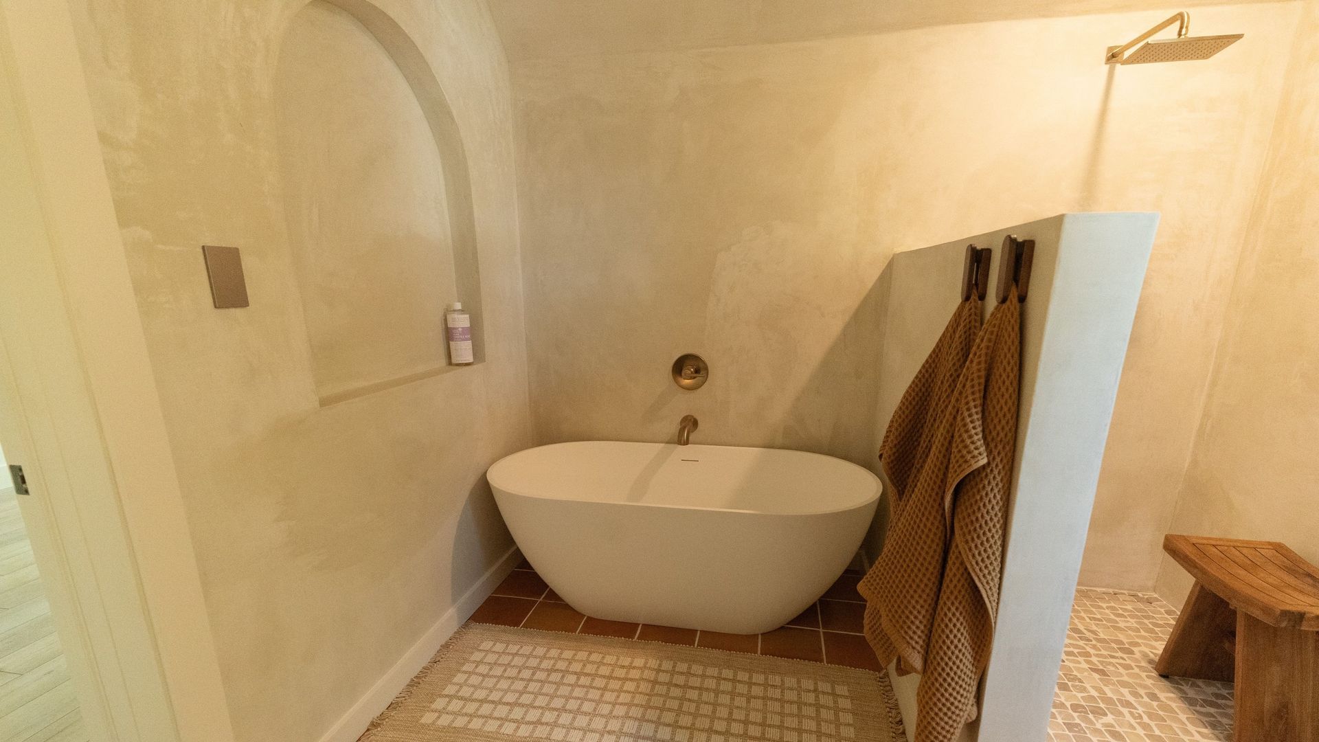 Bathroom with oval tub, shower area, and rustic wooden stool. Neutral walls, mosaic tile floor, and hanging towels.