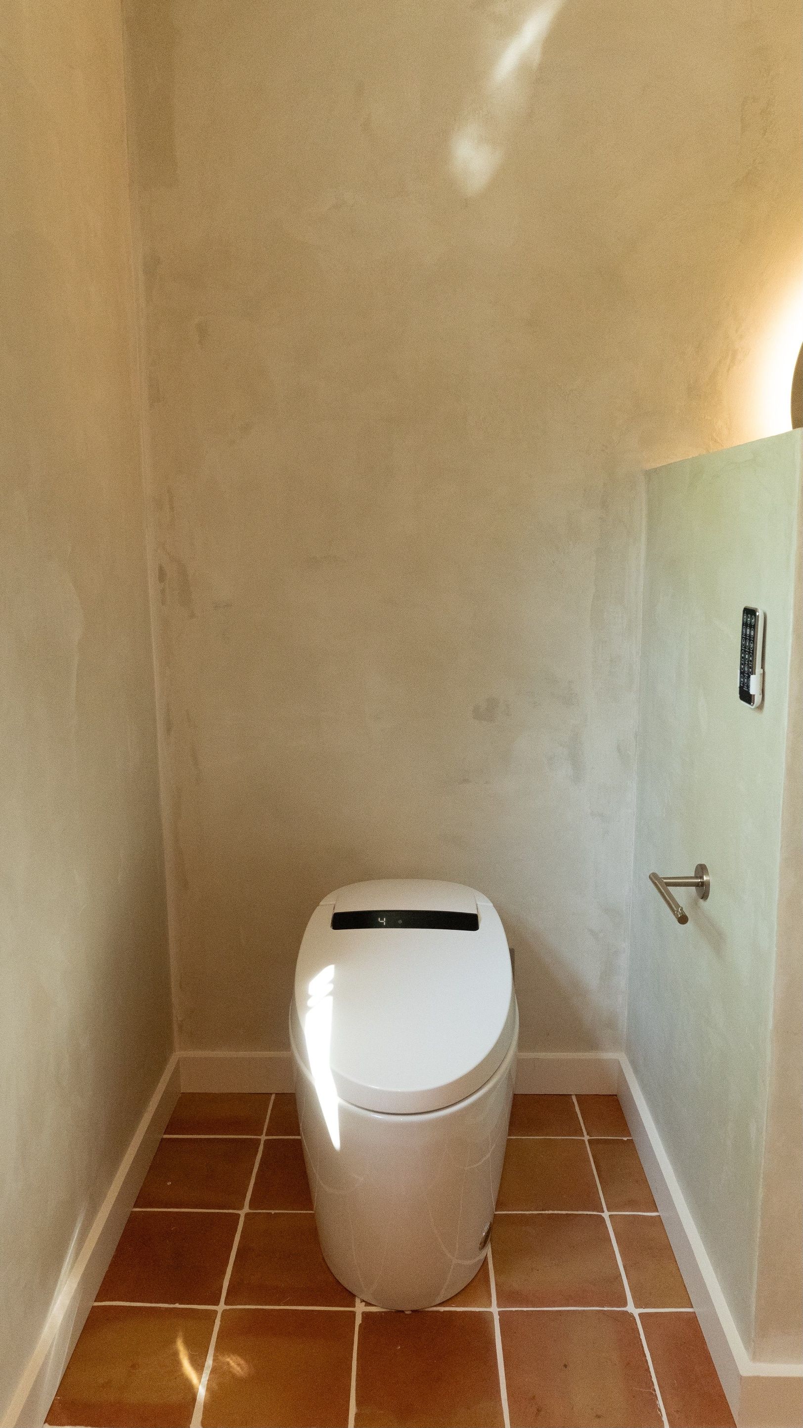 Toilet in a small bathroom with textured beige walls, red tile floor. Sunlight streams in.