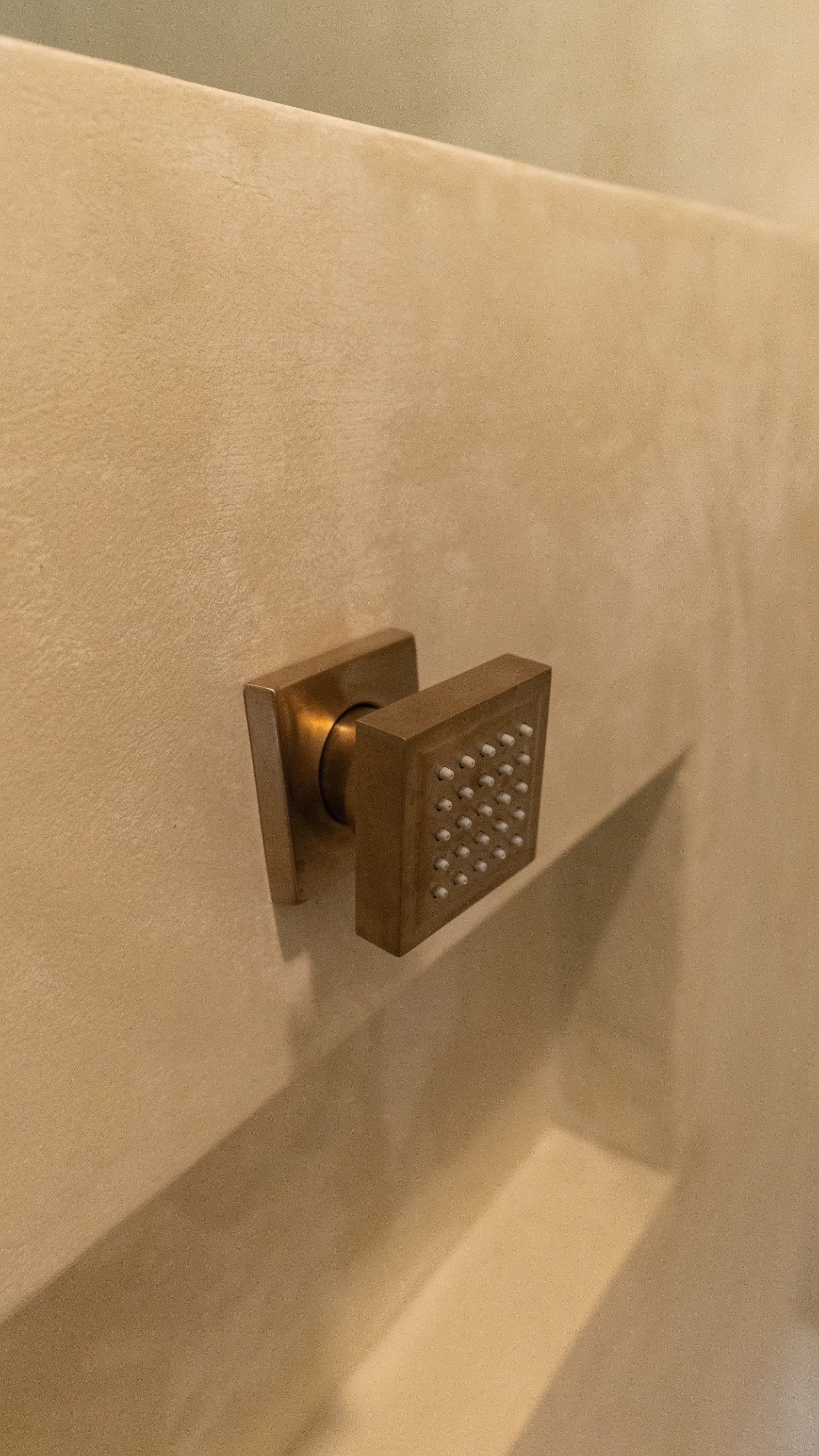 Bronze showerhead with a textured, square design mounted on a light beige wall.
