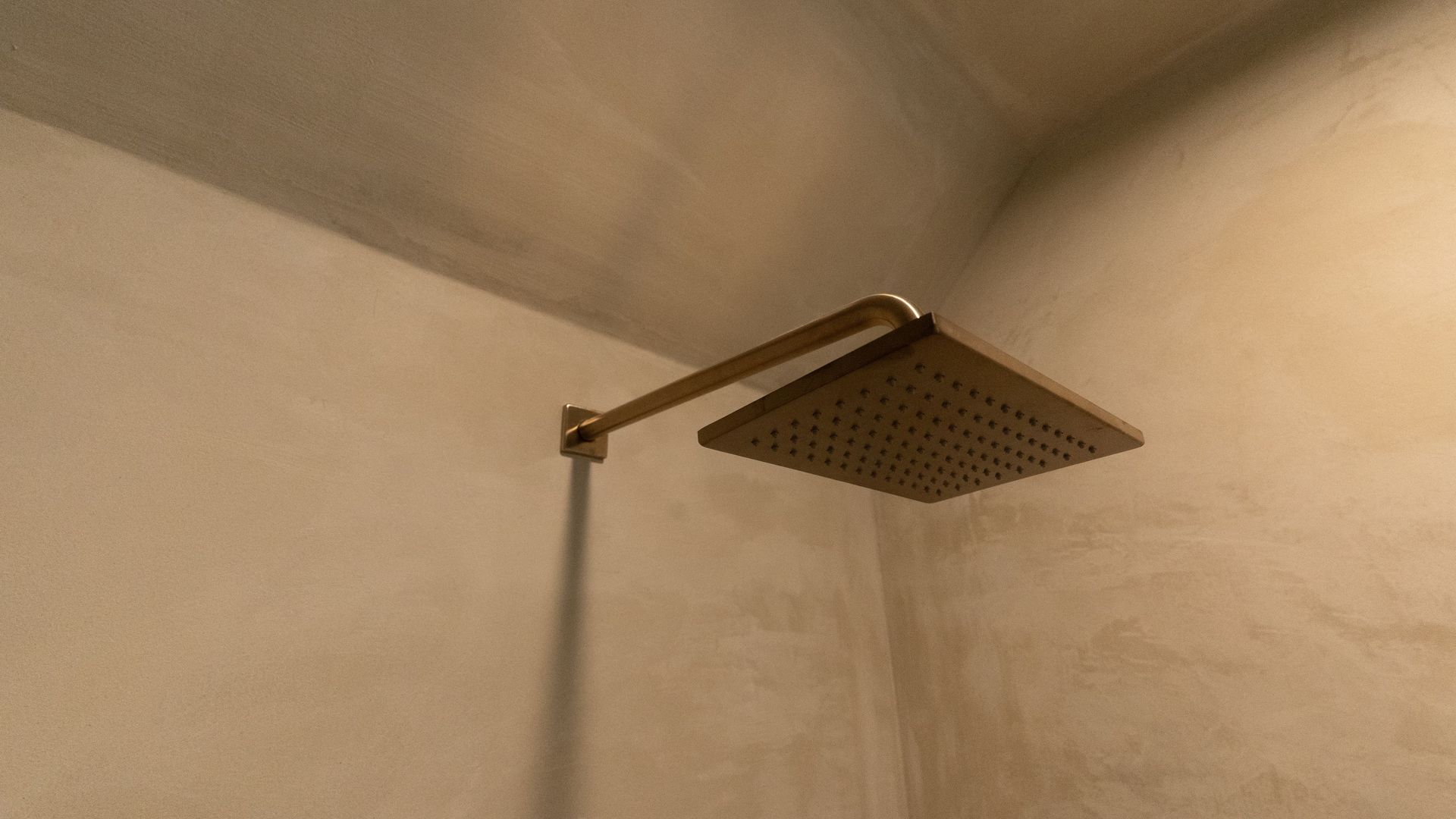 Square, brass showerhead mounted on a light-colored, textured wall.