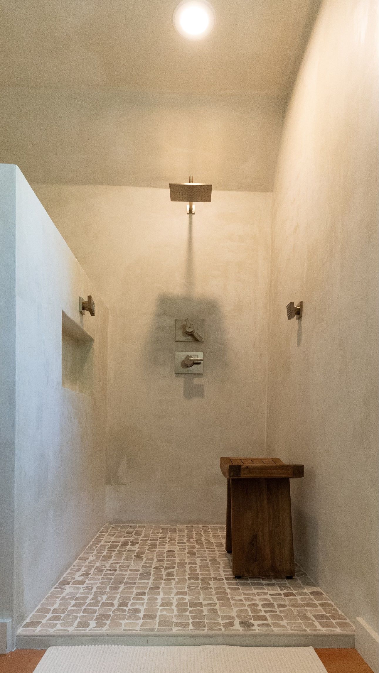 Shower with brick floor, wood bench, showerhead, and wall-mounted faucet; beige walls; bright ceiling light.