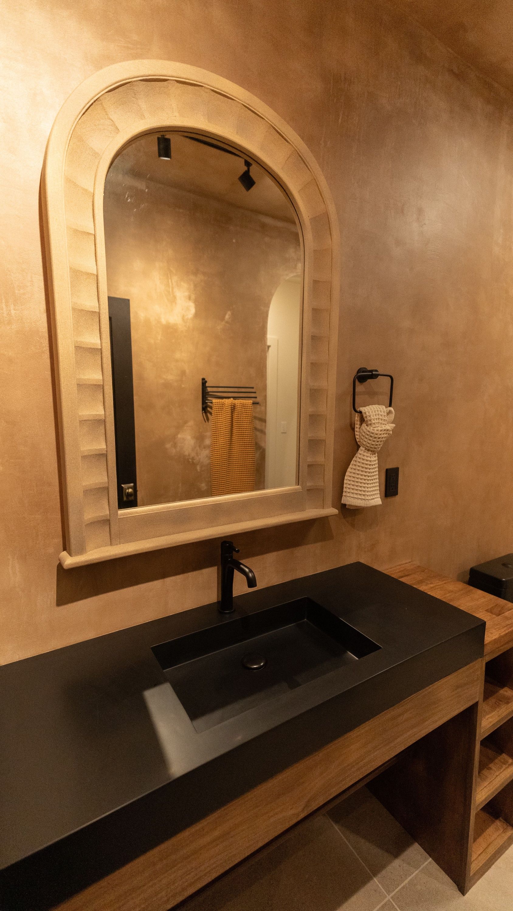 Bathroom with black sink, wooden vanity, arched mirror, and textured tan walls.