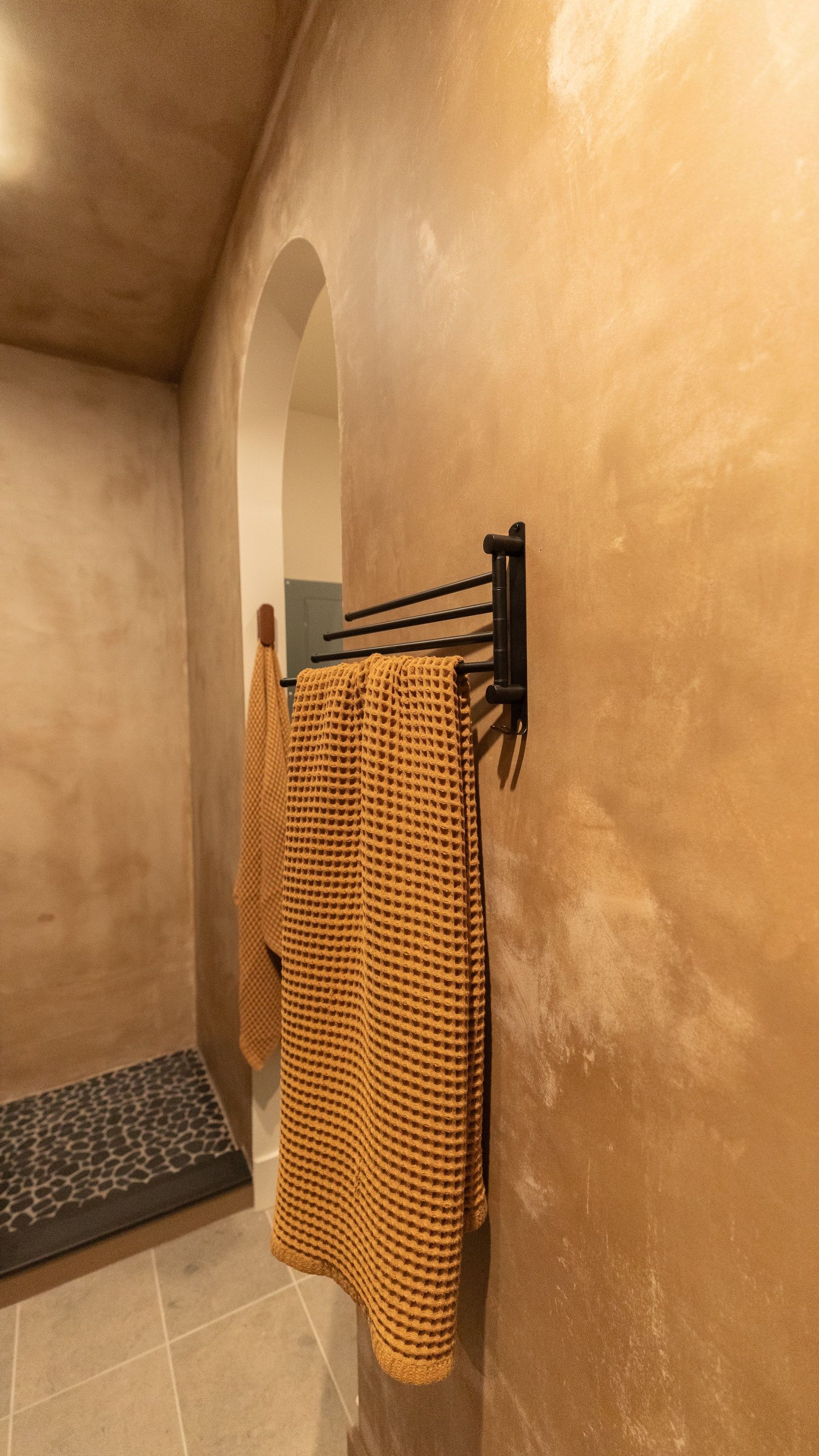 A black towel rack on a beige wall holds a golden-brown towel in a bathroom with an arched alcove.