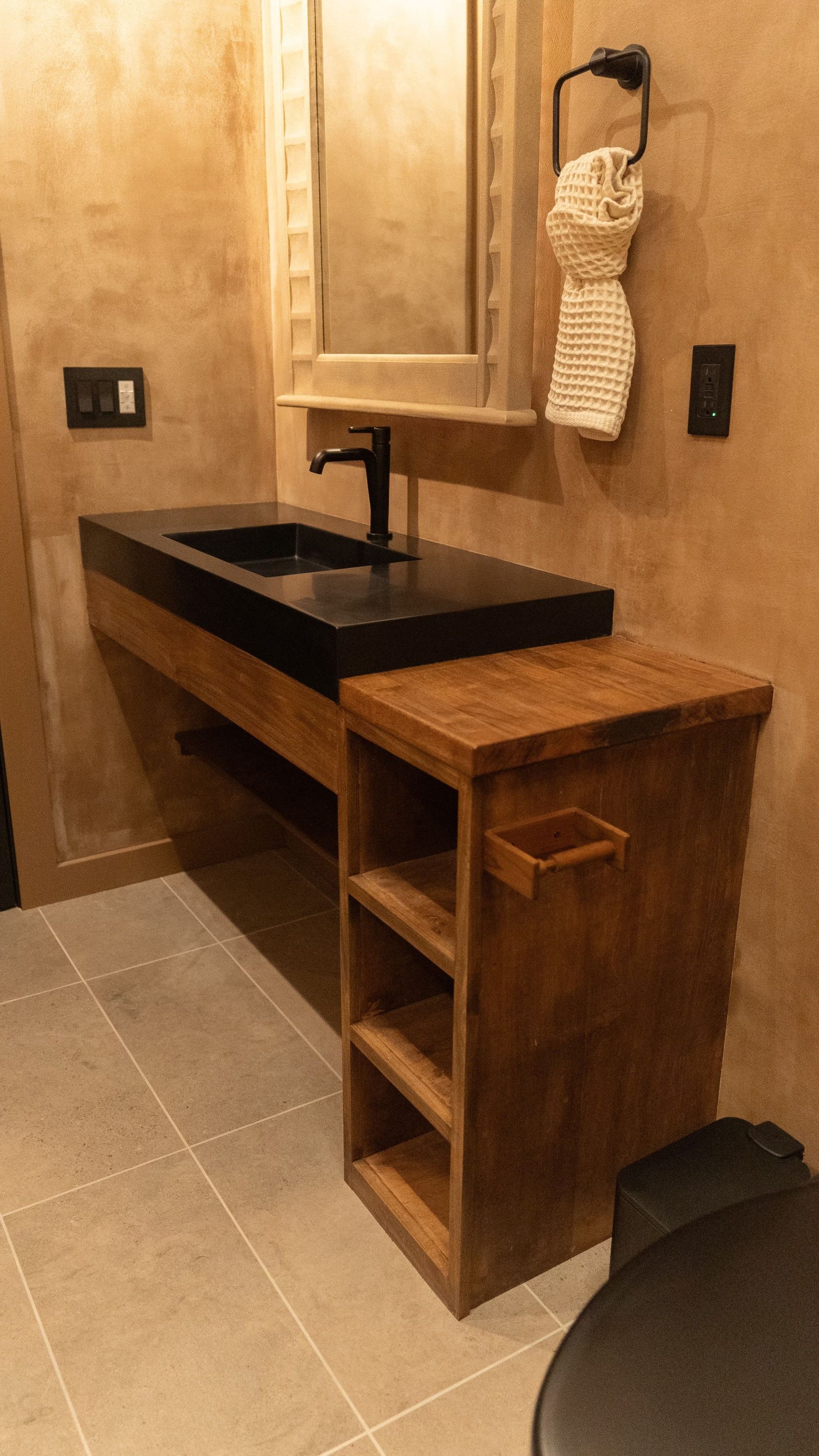 Bathroom with a black sink and faucet. A wooden cabinet with open shelves sits next to the toilet. Beige tile floor.