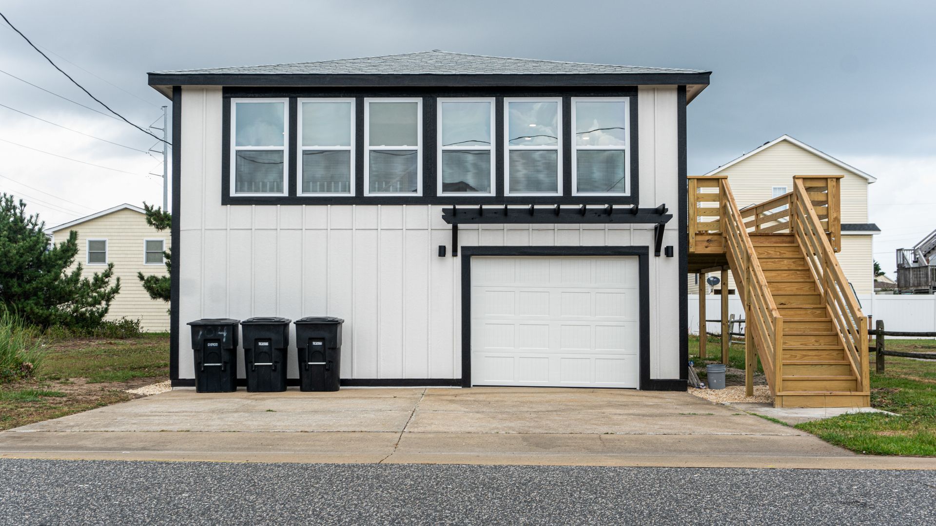 White two-story beach house with black trim, garage, and wooden stairs under a cloudy sky.