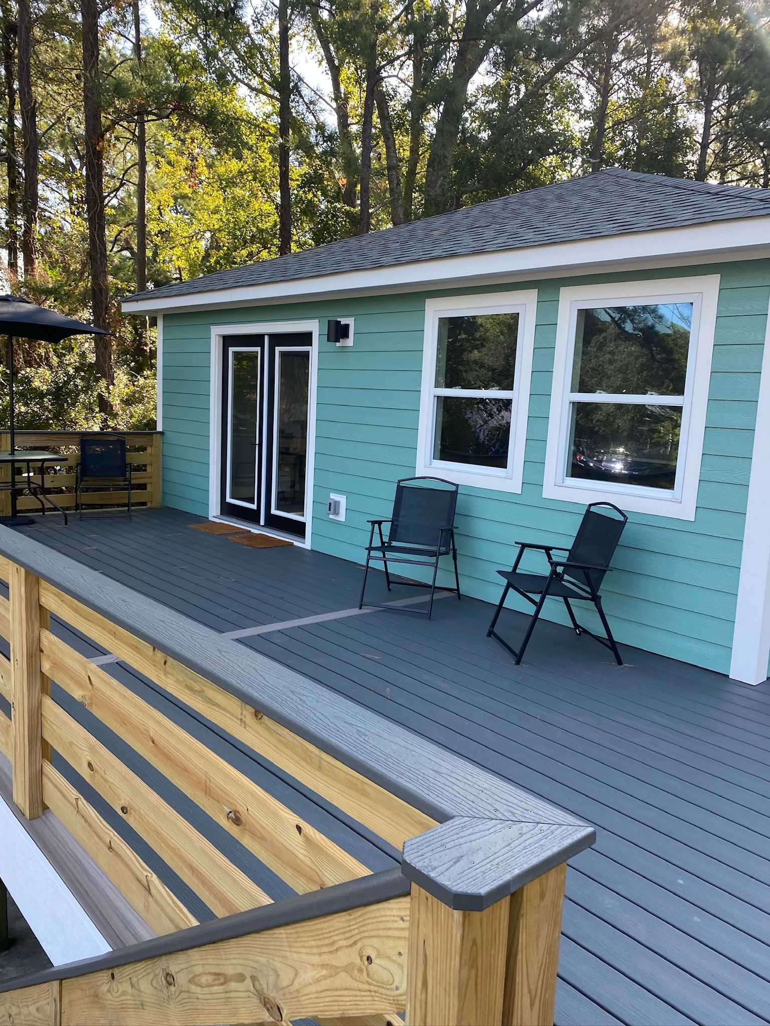 Teal house with deck; two chairs outside French doors; gray deck, wood railing, trees in the background.