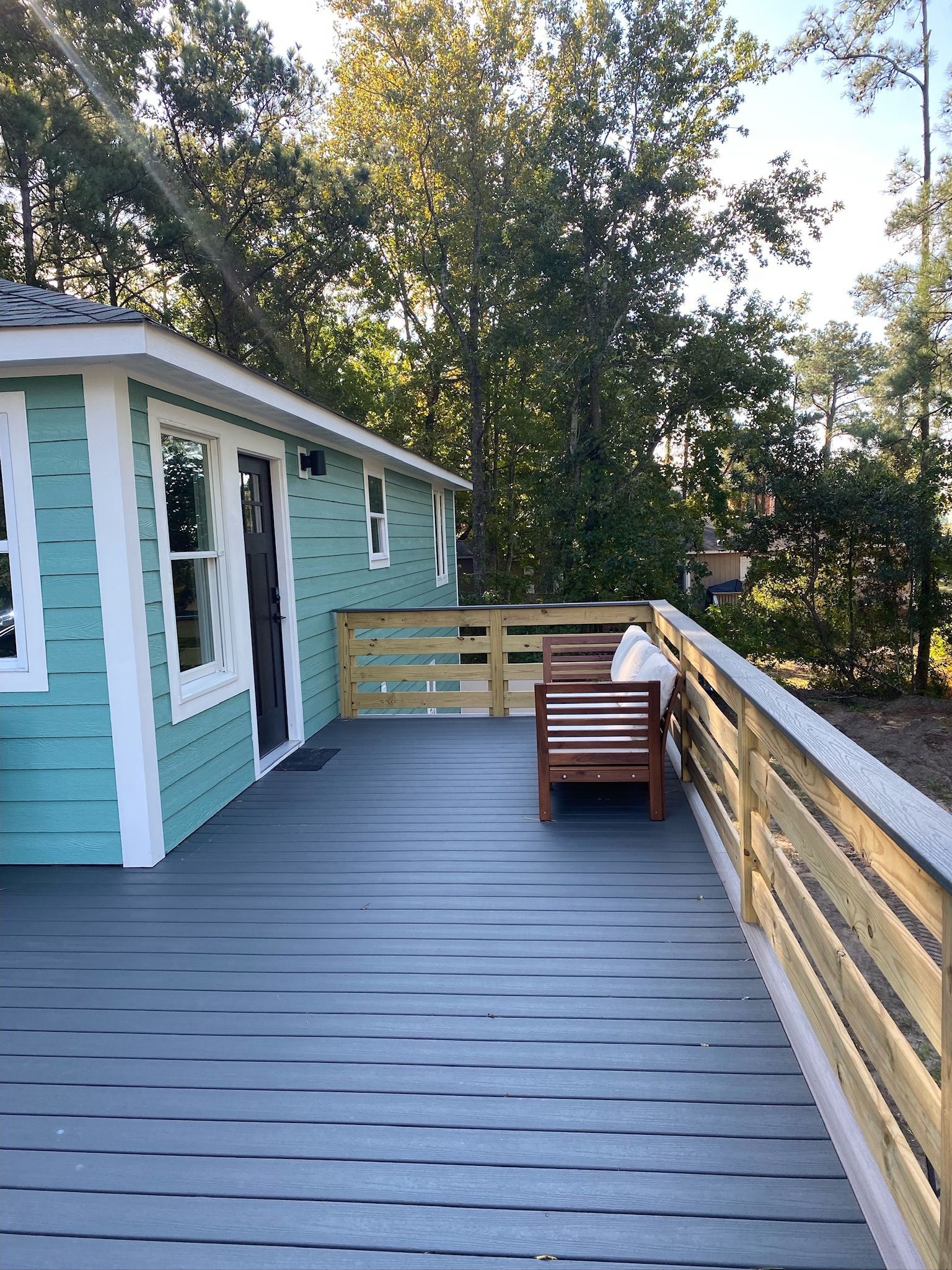 Teal-colored house with a gray deck, wooden chair, and wood railing surrounded by trees.