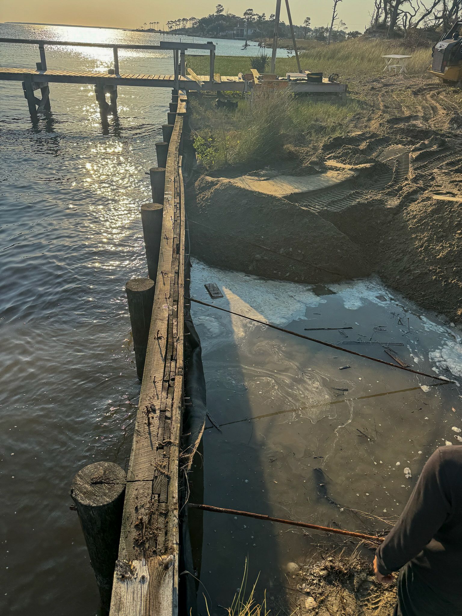 Wooden pier at a murky shore with a person in the corner; sunlight reflects on the water.