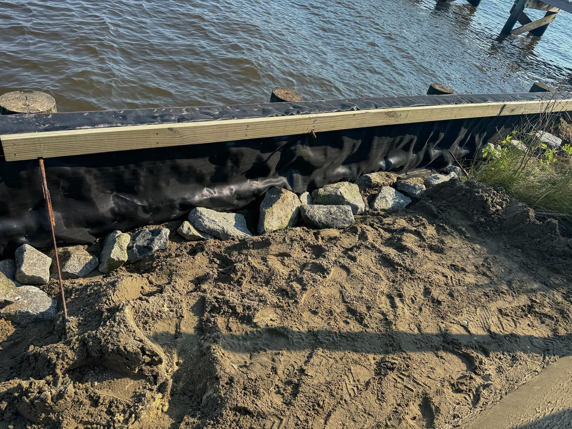 A waterside construction. Dark fabric covers rocks, under a wooden beam. Soil is in the foreground.