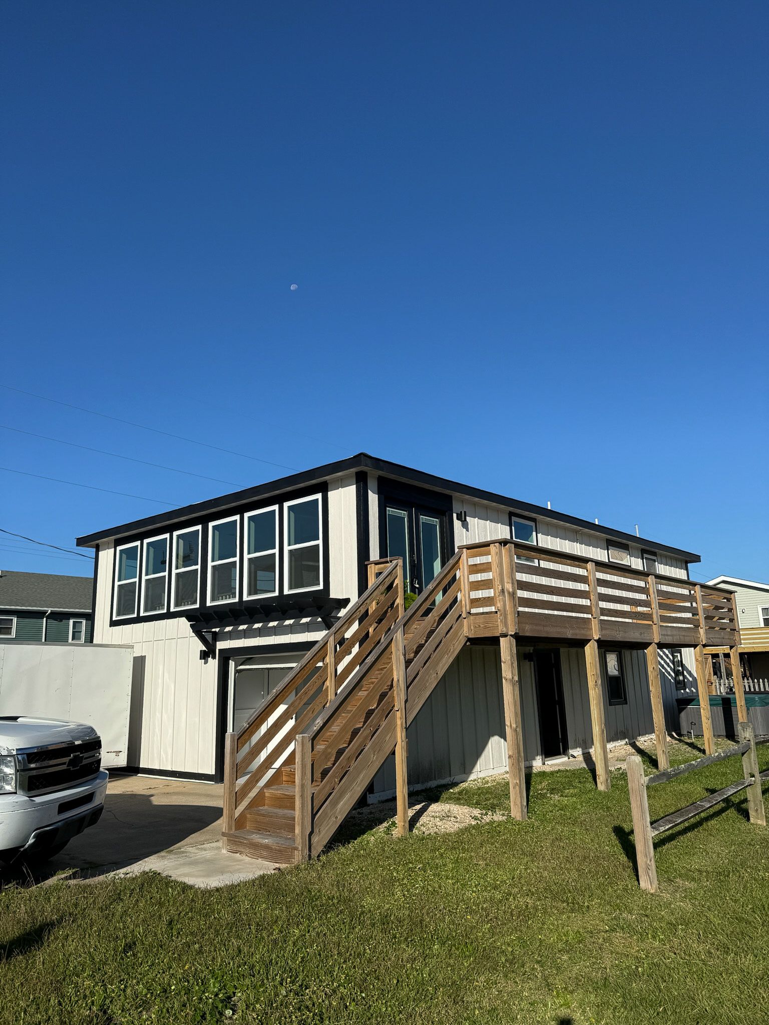 Two-story beach house with wooden deck, stairs, and windows, white and black exterior, on green grass.