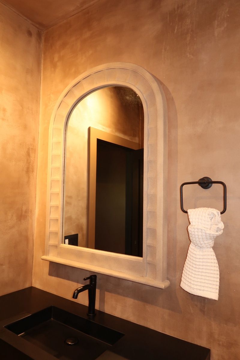 Arched mirror with a black sink and faucet in a bathroom with textured tan walls. White towel on black towel rack.