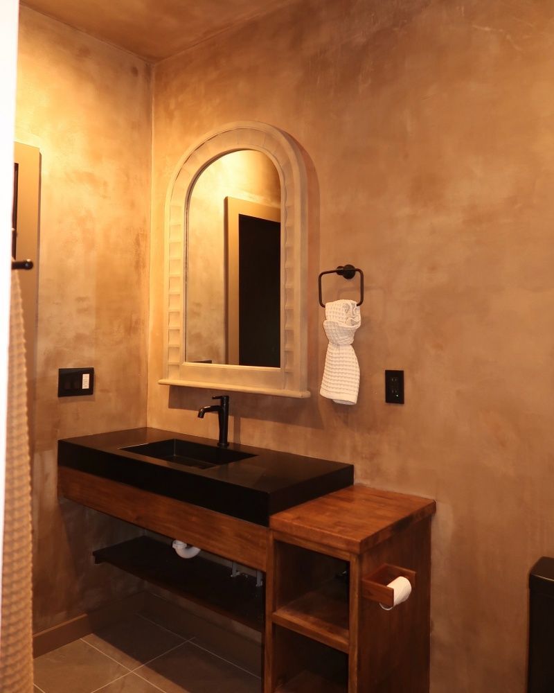 Bathroom with dark sink, wooden vanity, arched mirror, and textured brown walls.