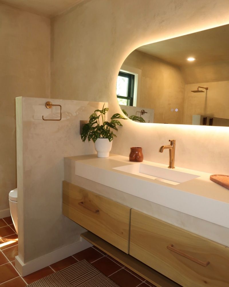 Modern bathroom with concrete countertop, wood cabinets, arched mirror with backlighting, and gold fixtures.