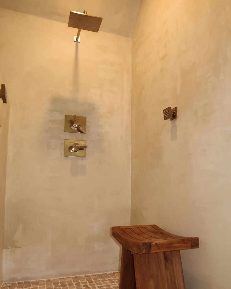 Shower stall with tan walls, brass fixtures, wooden bench, and mosaic tile floor.