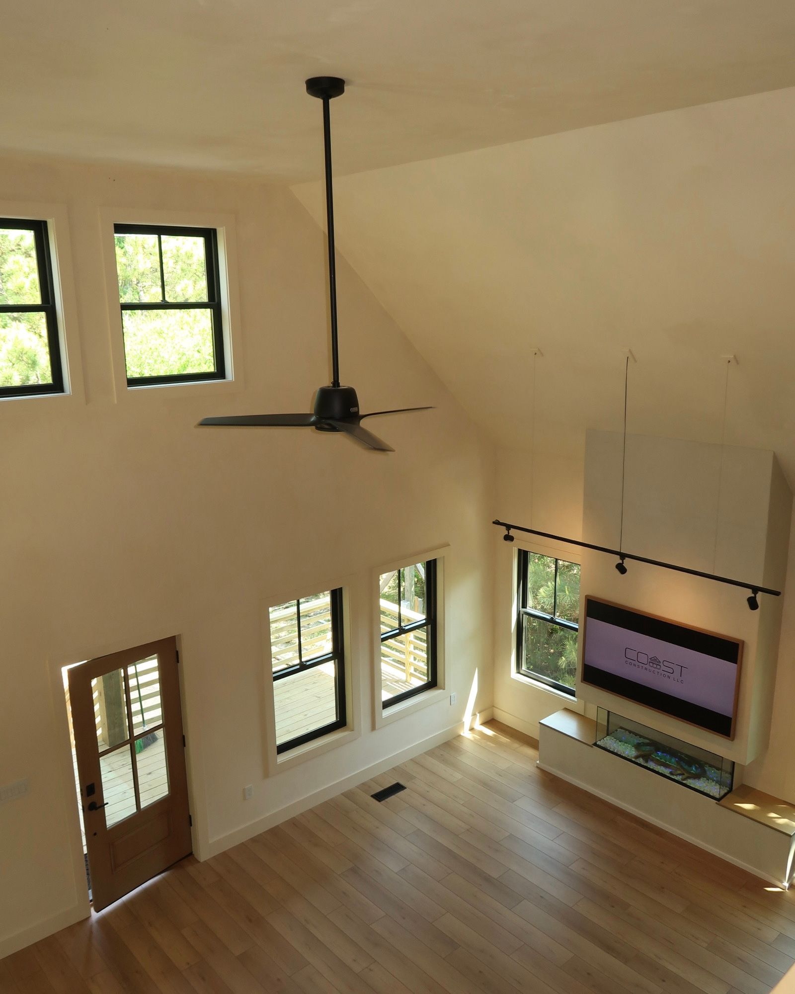 High-ceiling living room with black-framed windows, a ceiling fan, hardwood floors, and a TV mounted on a white cabinet.