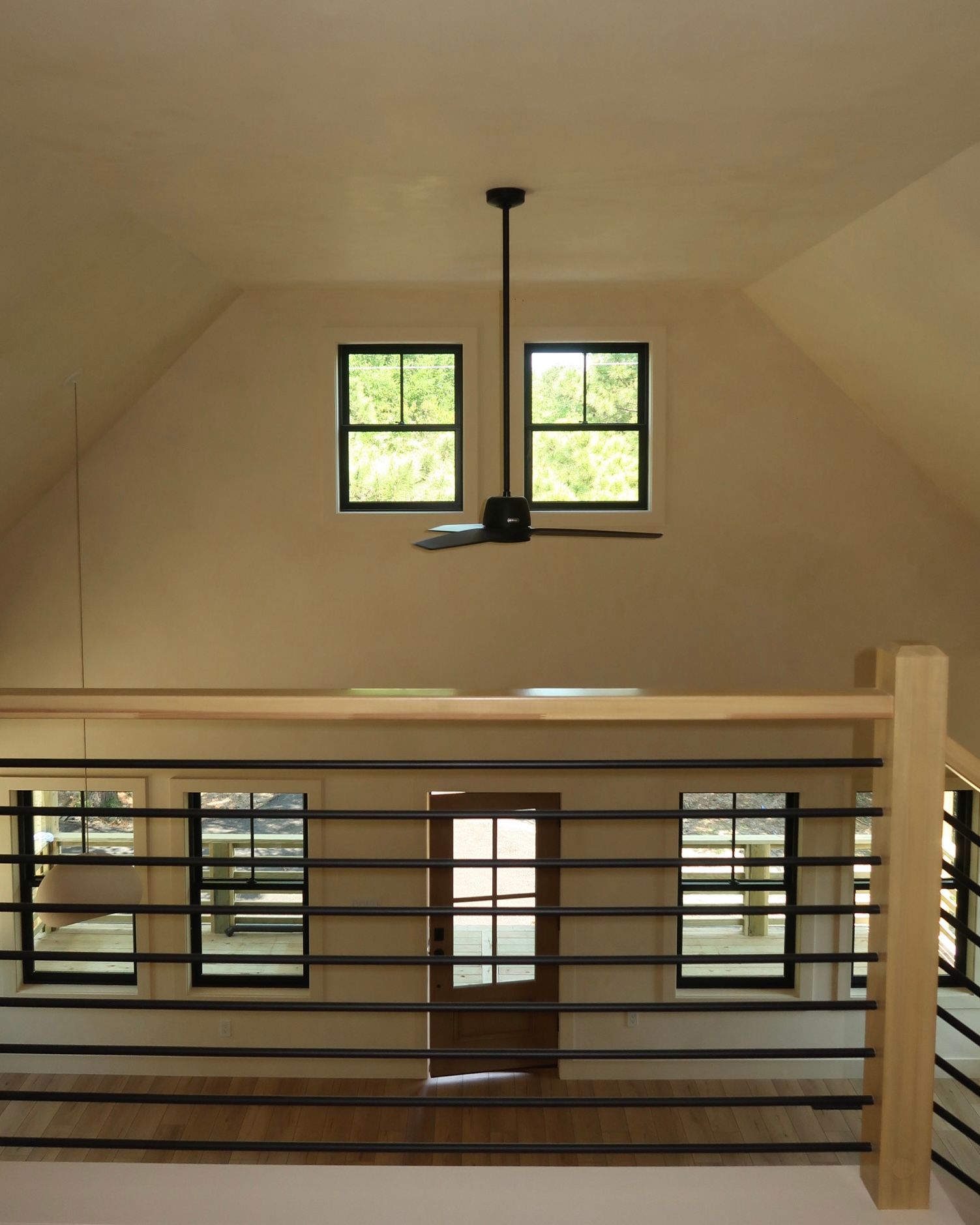 Interior view of a loft with windows, a door, and a black ceiling fan. Beige walls and light wood railings.
