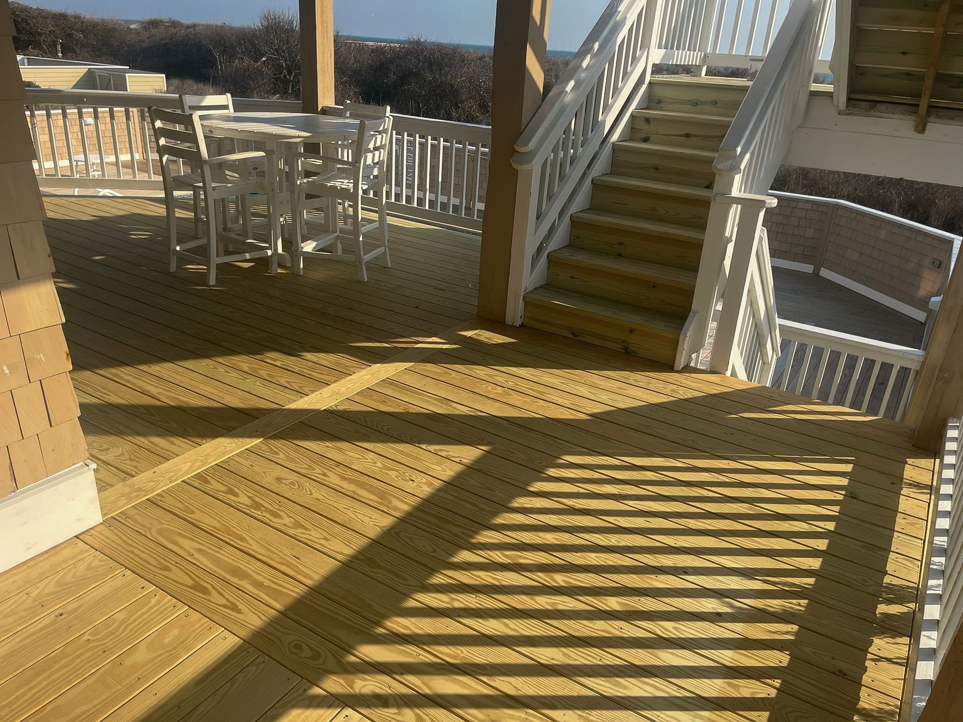 Wooden deck with stairs, a table, and chairs. Bright sunlight casts shadows.