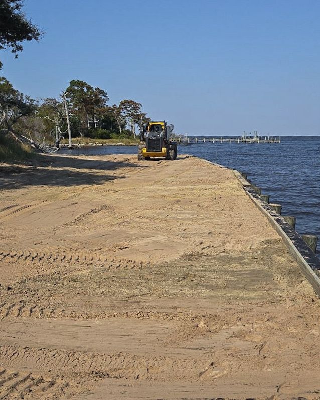 A yellow skid steer on a sandy beach; water and a pier are in the background.