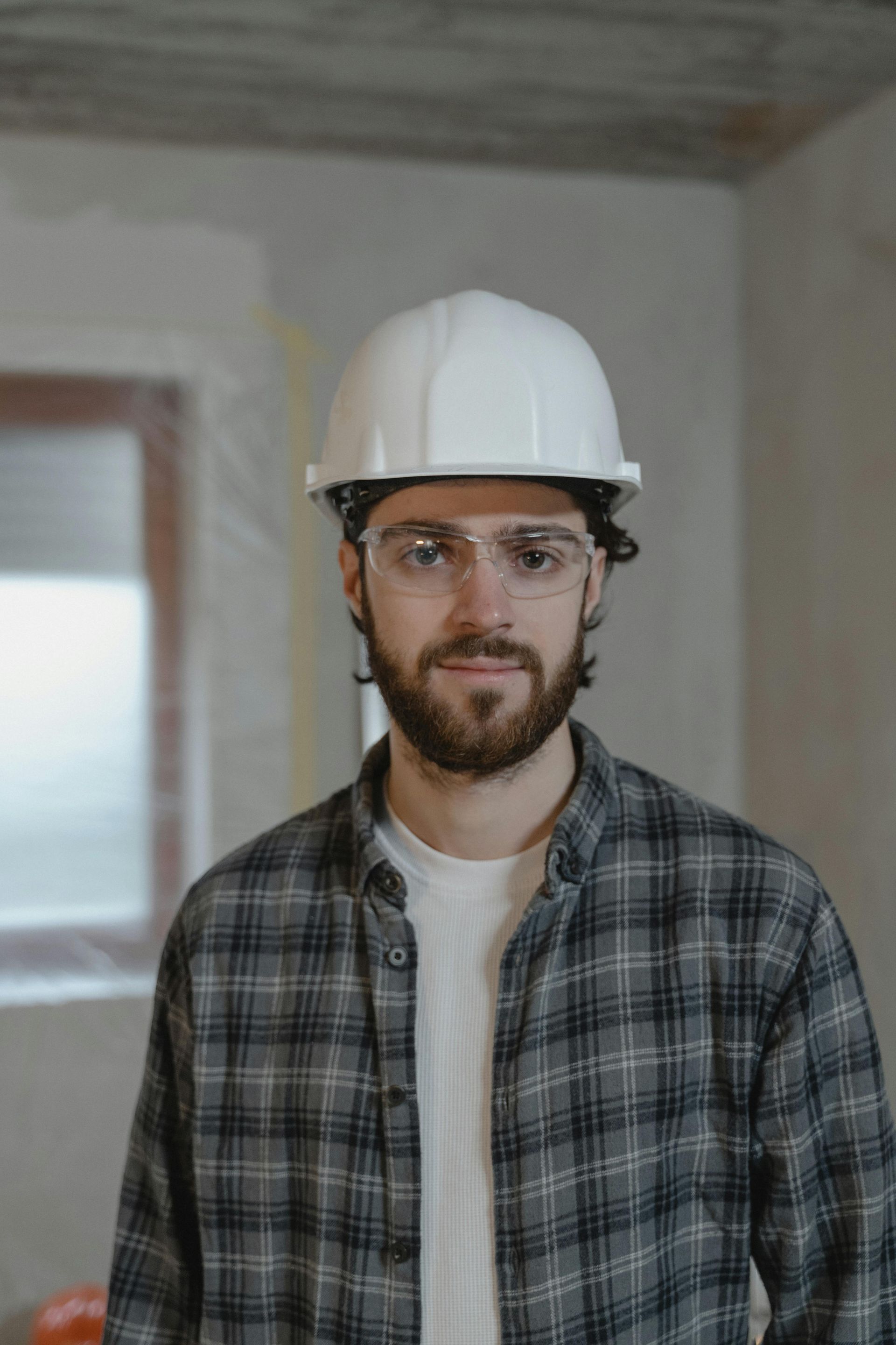 Man with beard wearing a white hard hat and safety glasses, in a room under construction.