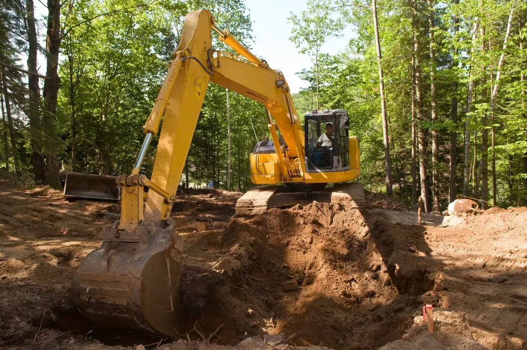 Yellow excavator digging in dirt, surrounded by trees.