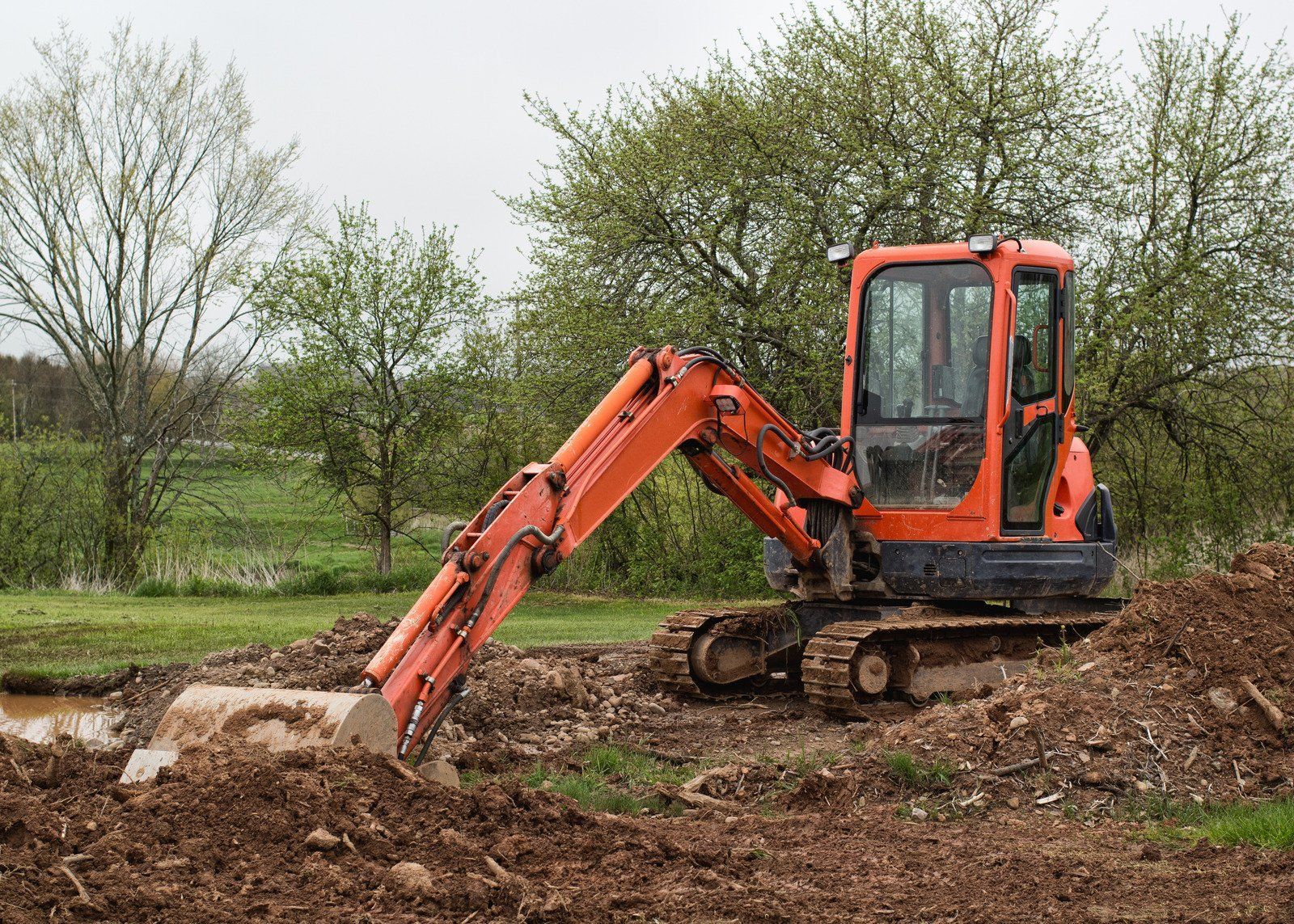 Orange excavator digging in muddy soil outdoors. Trees and cloudy sky in the background.